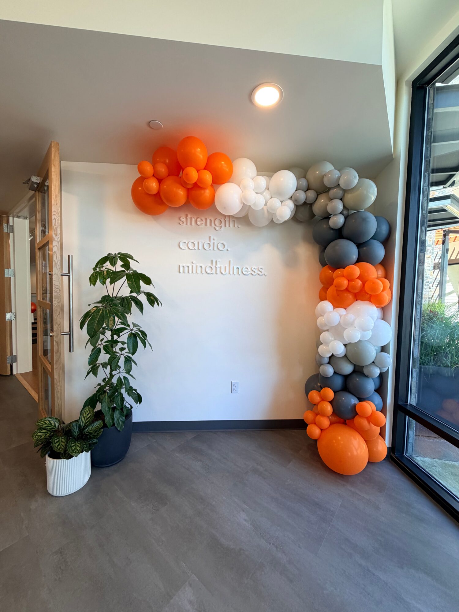 Balloon arrangement with orange, white, gray, and black balloons near a window, with a potted plant and wall text.