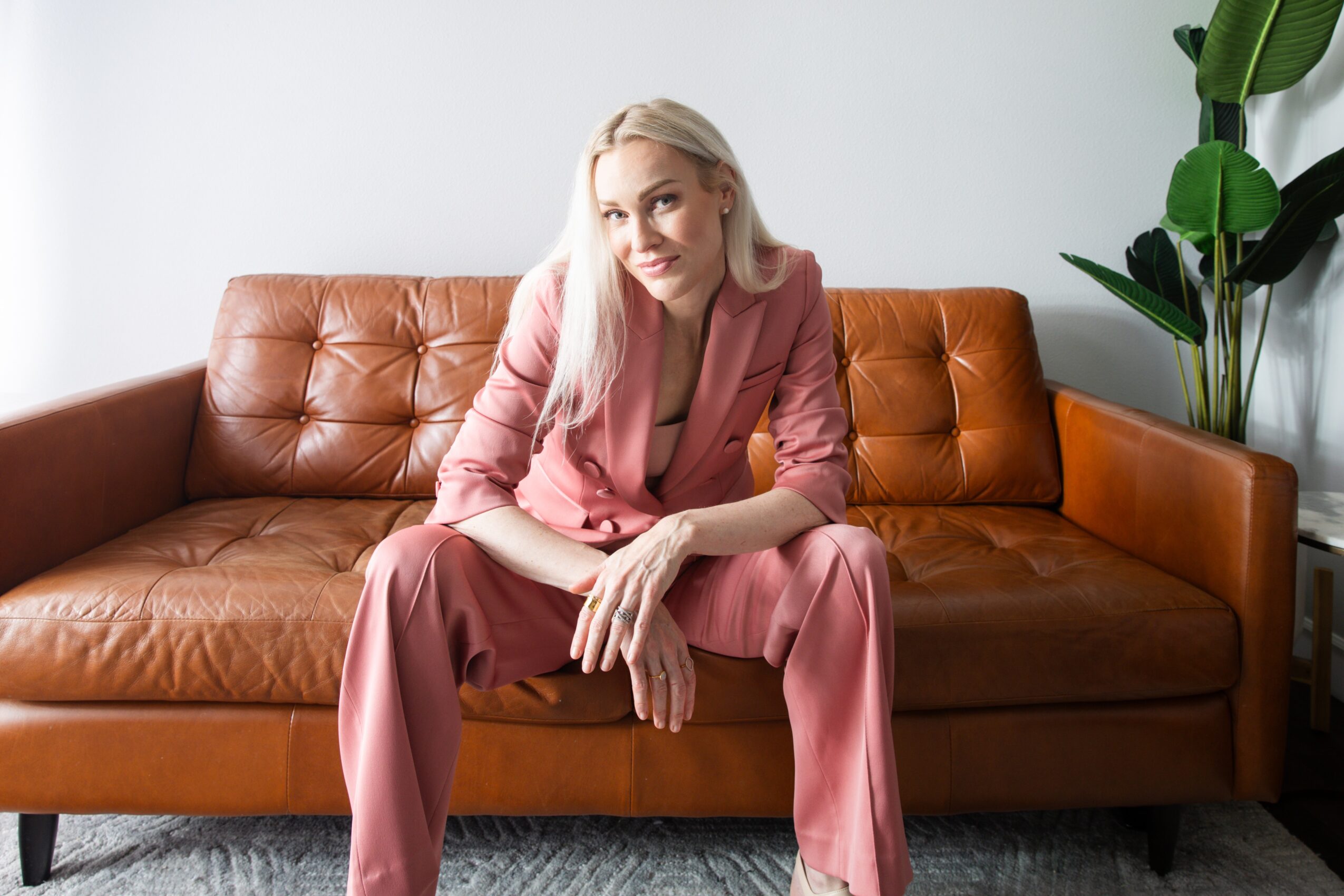 Woman with blonde hair in pink suit sitting on a brown leather sofa, with a green plant nearby.