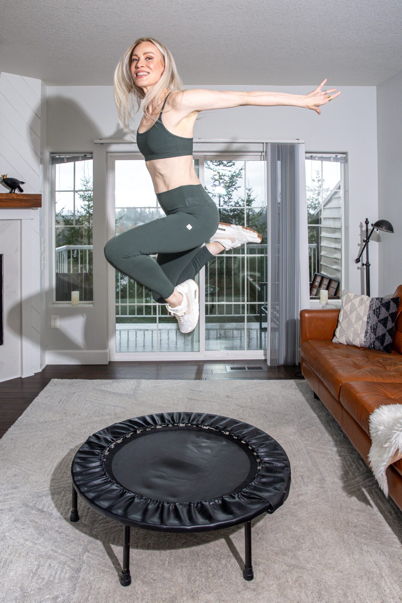 Young woman jumping on a trampoline indoors, smiling, with a living room background including a window and furniture.