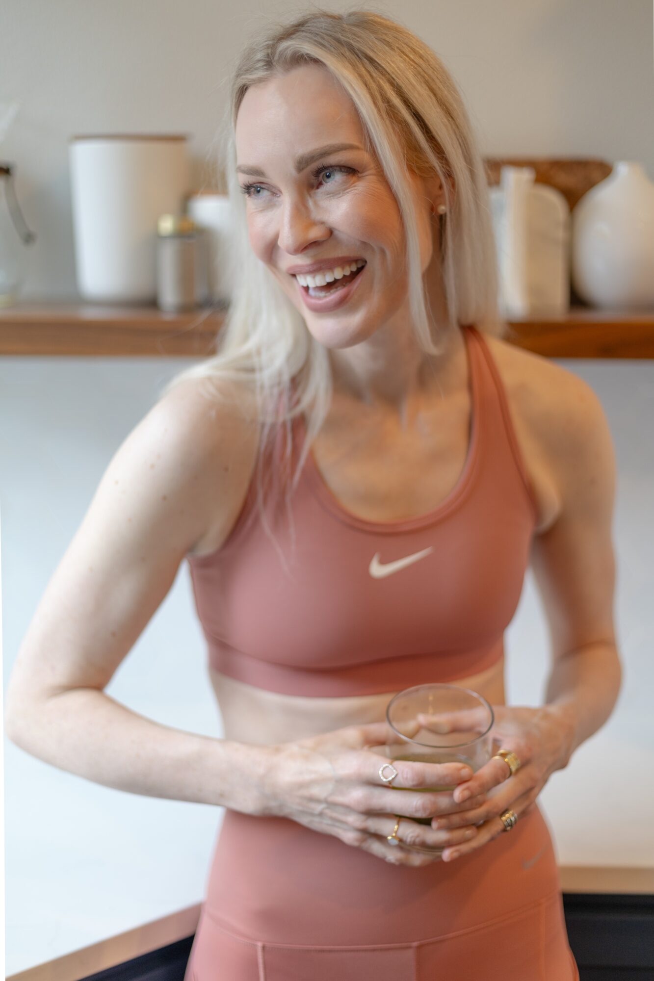 Smiling woman in workout clothes holding a glass, standing in a kitchen with jars and containers in the background.