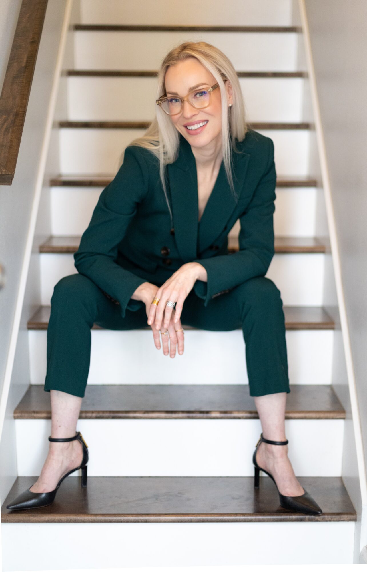 Woman with blonde hair, glasses, in a dark green suit, sitting on stairs, smiling, with crossed legs and heels.
