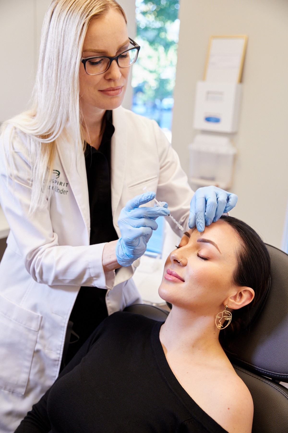 Medical professional in white coat and gloves administers treatment to a woman with closed eyes, seated in a clinic.