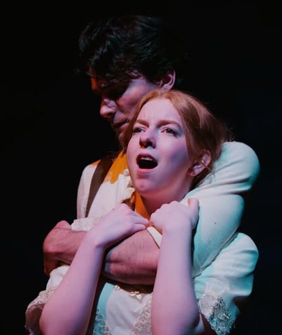 Two people in a dramatic scene, one with dark hair and the other with red hair, embracing each other with intense expressions.