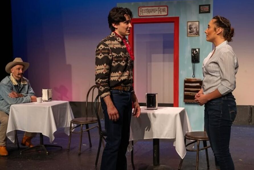 Two people stand facing each other in a room with a table and chairs, a person sits at the table, and a door is in the background.