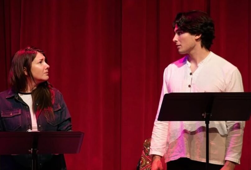 Two people standing on stage with red curtains, facing each other, one with long dark hair and the other with dark curly hair, both near music stands.