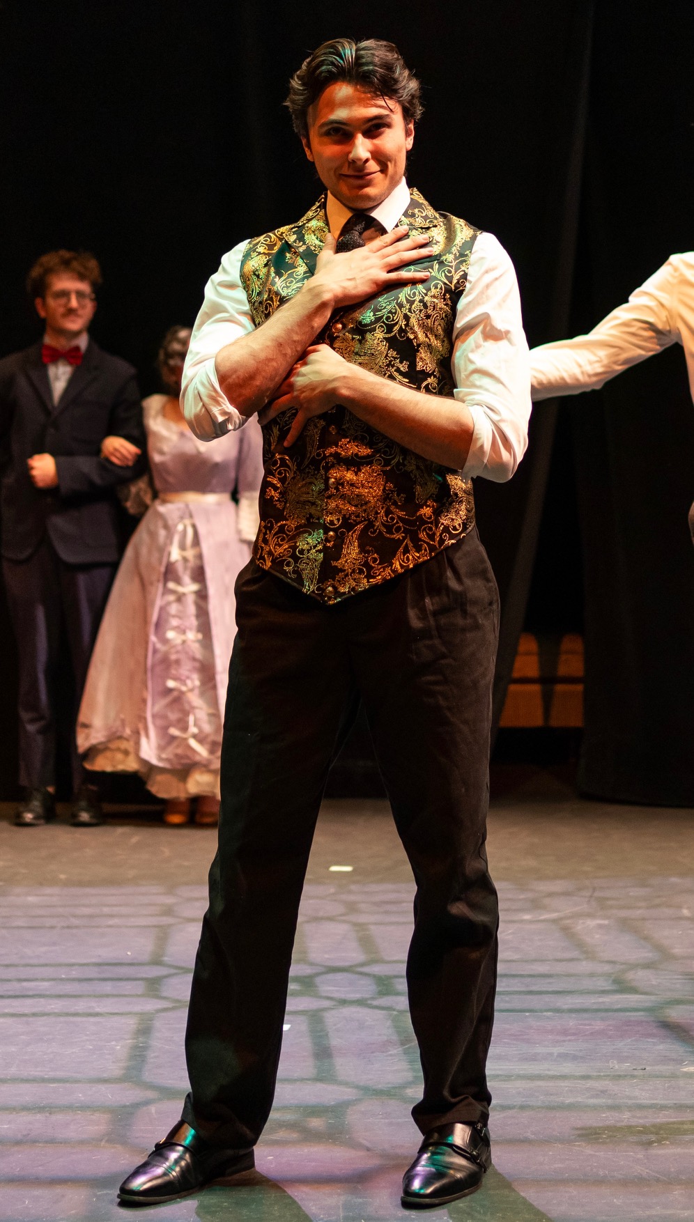 Person standing on stage with hand on chest, wearing a patterned vest and black pants, with others in background.