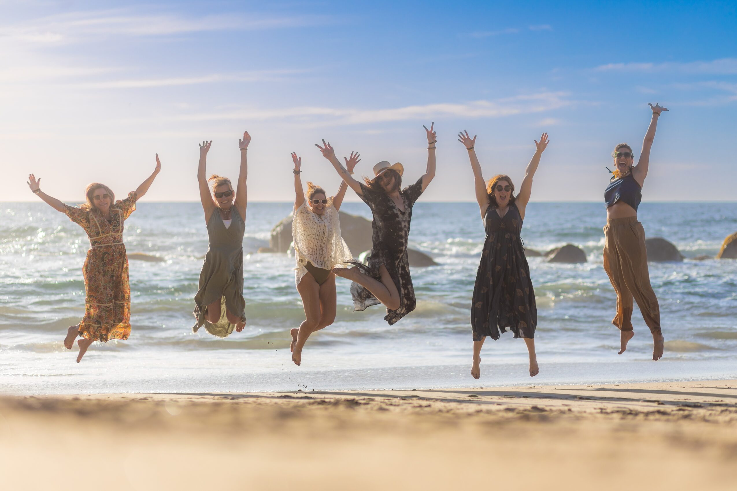 Six people jumping on the beach with arms raised, ocean waves in the background, sunny sky, and rocks in the water.