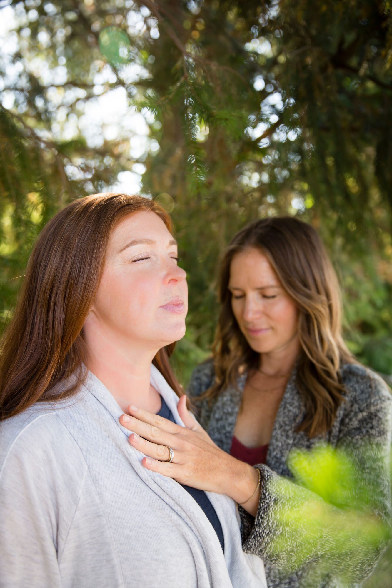 Two women outdoors, one with eyes closed, the other touching her chest, surrounded by trees and sunlight, peaceful scene.