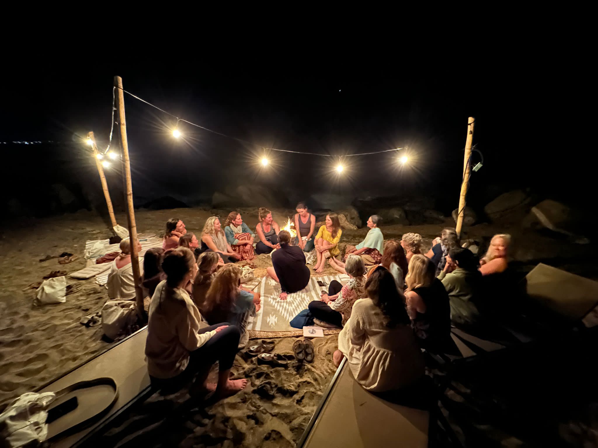 Group of people sitting around a table outdoors at night, illuminated by string lights, on a sandy surface.