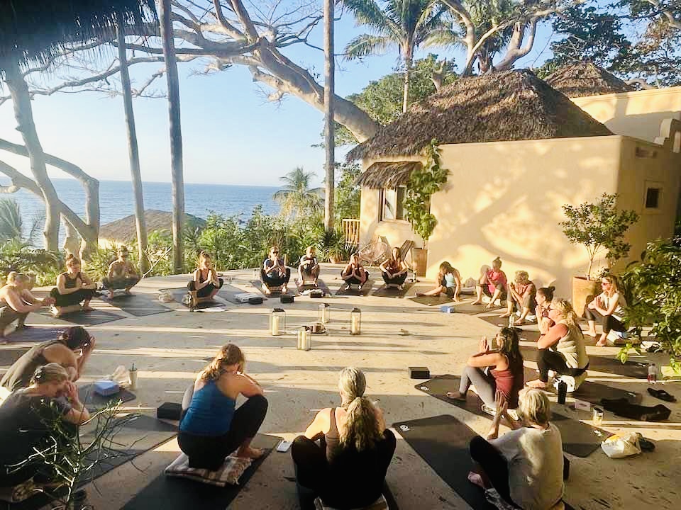 Group of people practicing yoga outdoors on a terrace with trees and ocean view in the background.