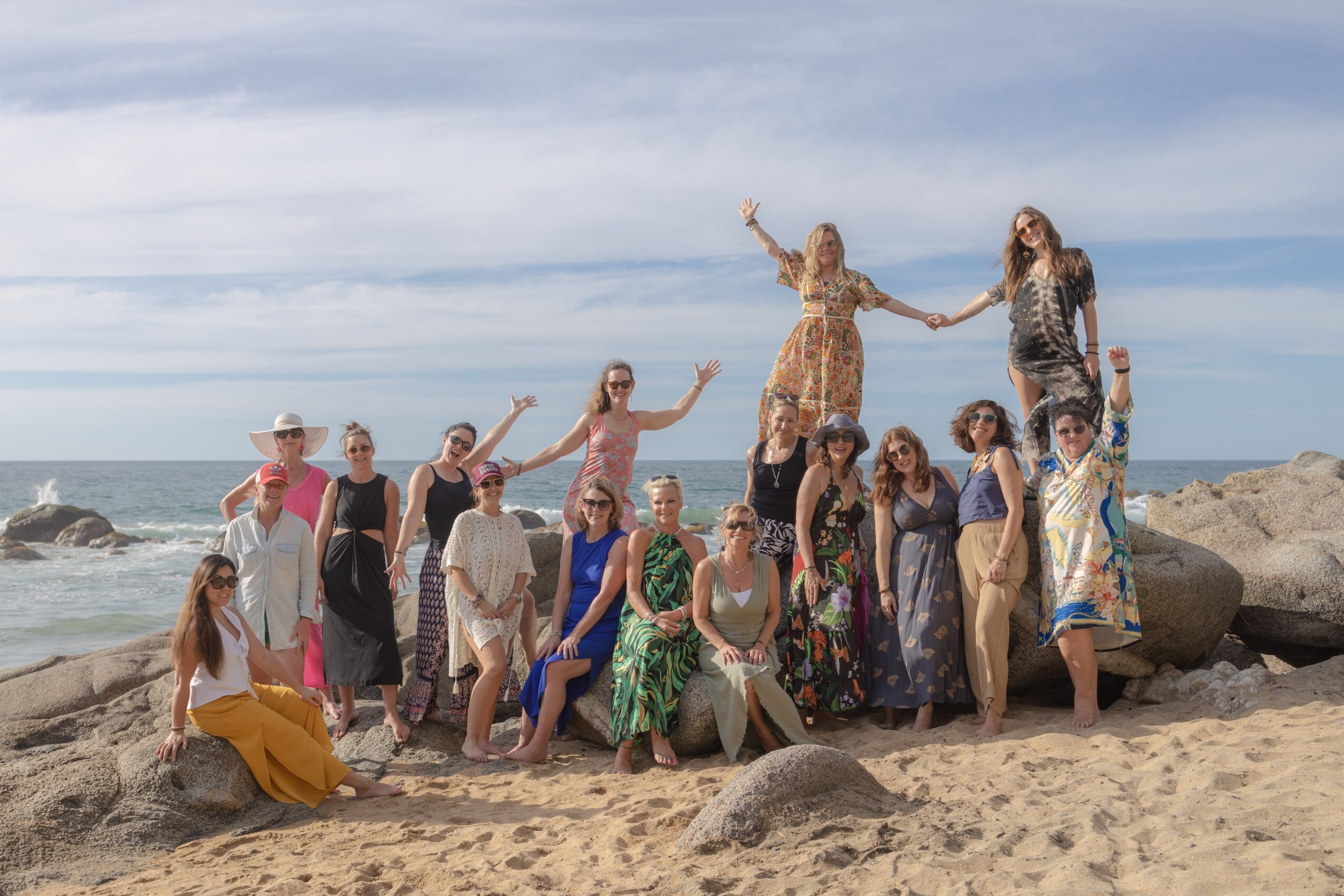 Group of women on a beach with rocks, some holding hands, under a partly cloudy sky.