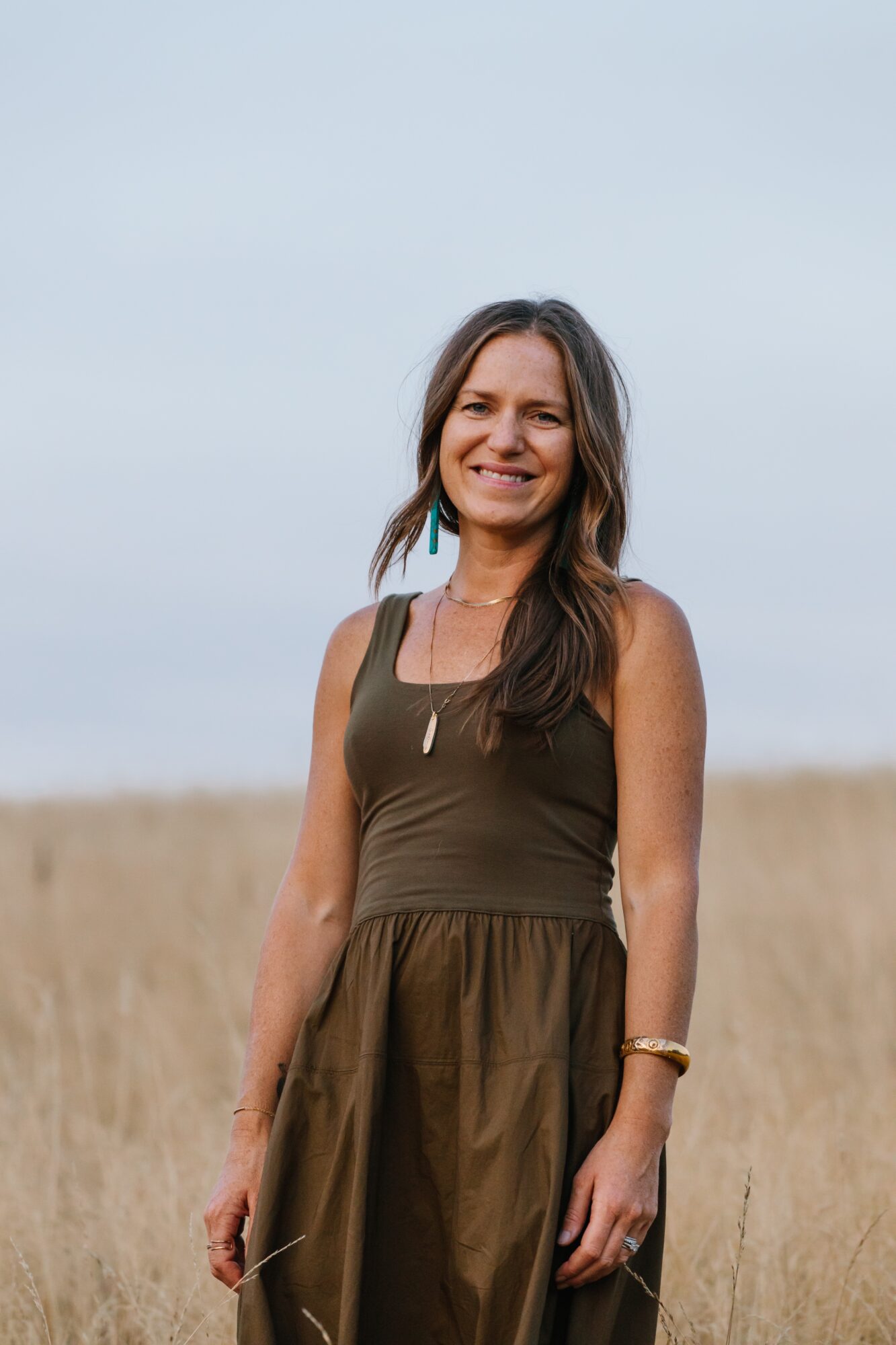Smiling woman standing outdoors in a field with a cloudy sky background, wearing a sleeveless dress and a bracelet.