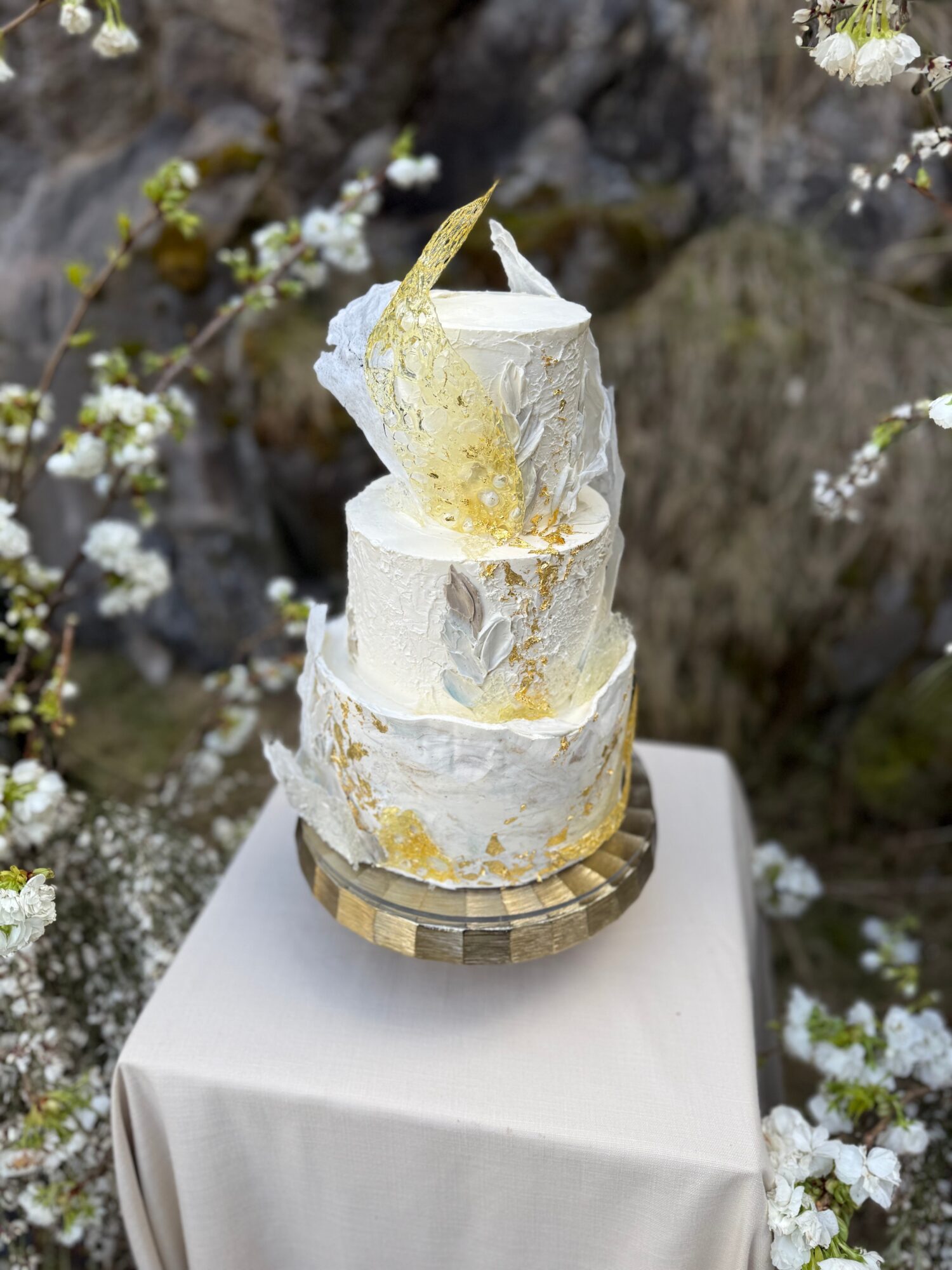Three-tiered white cake with gold accents, decorated with a gold shard, on a gold stand, outdoors with blooming branches