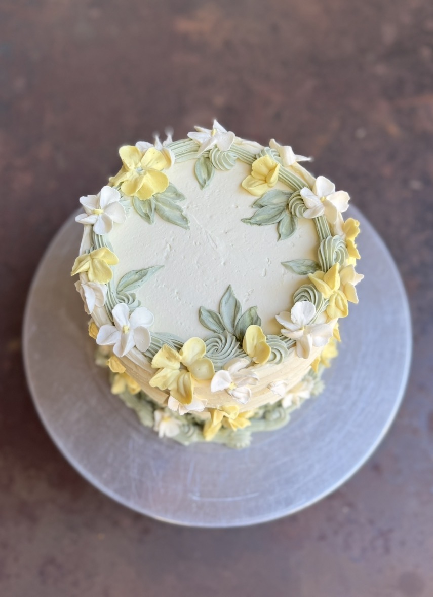 Small round cake decorated with yellow and white flowers and green leaves, on a silver cake board.