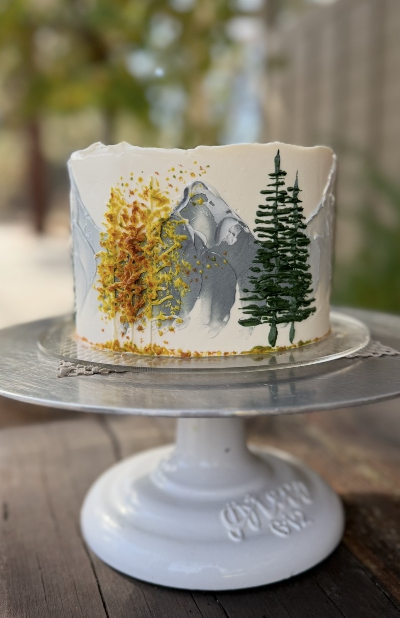 Cake with painted mountain, trees, and yellow foliage, on a white cake stand outdoors.