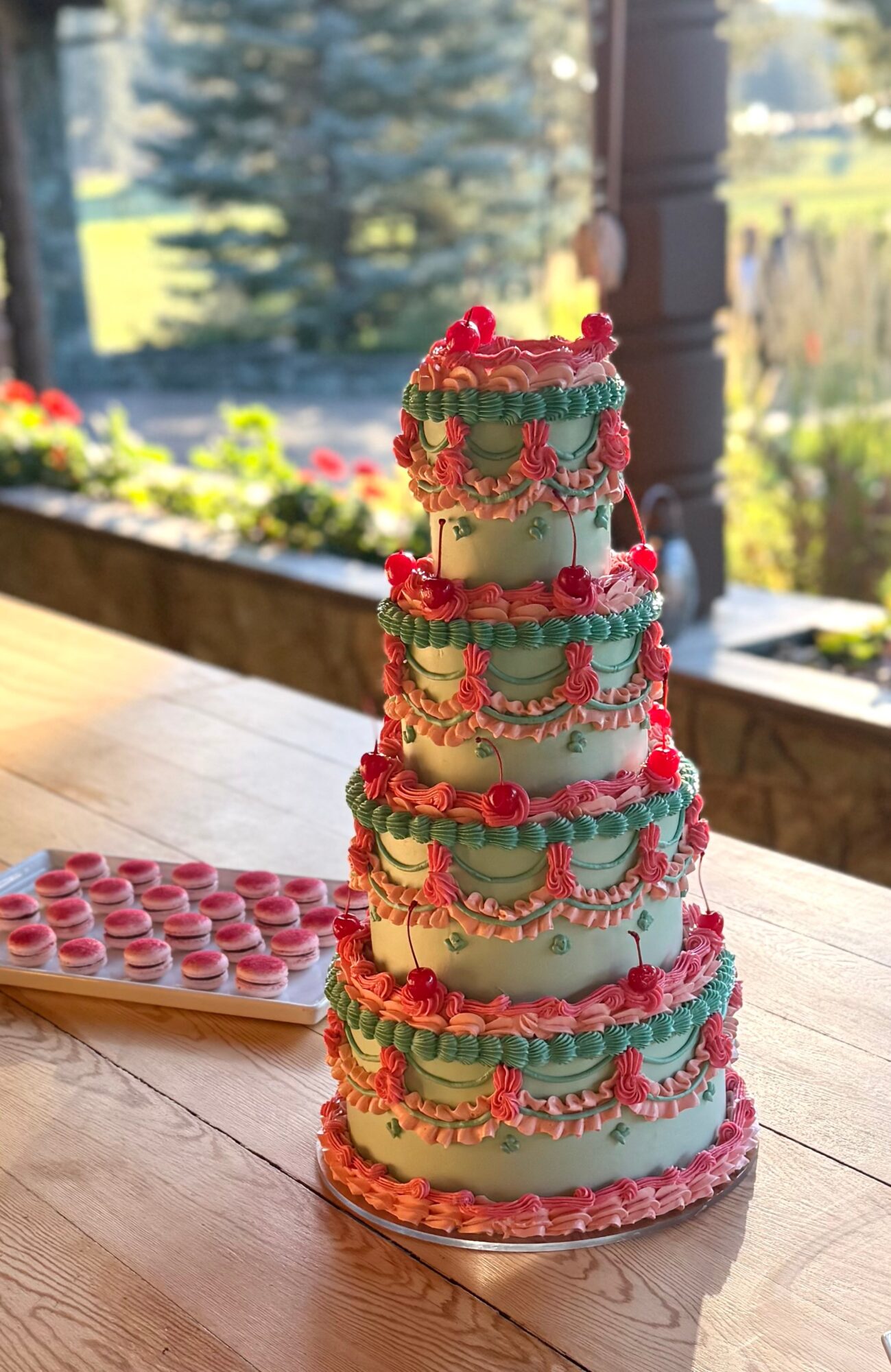 Four-tiered cake decorated with red and green icing, topped with cherries, on a wooden table with a window view.