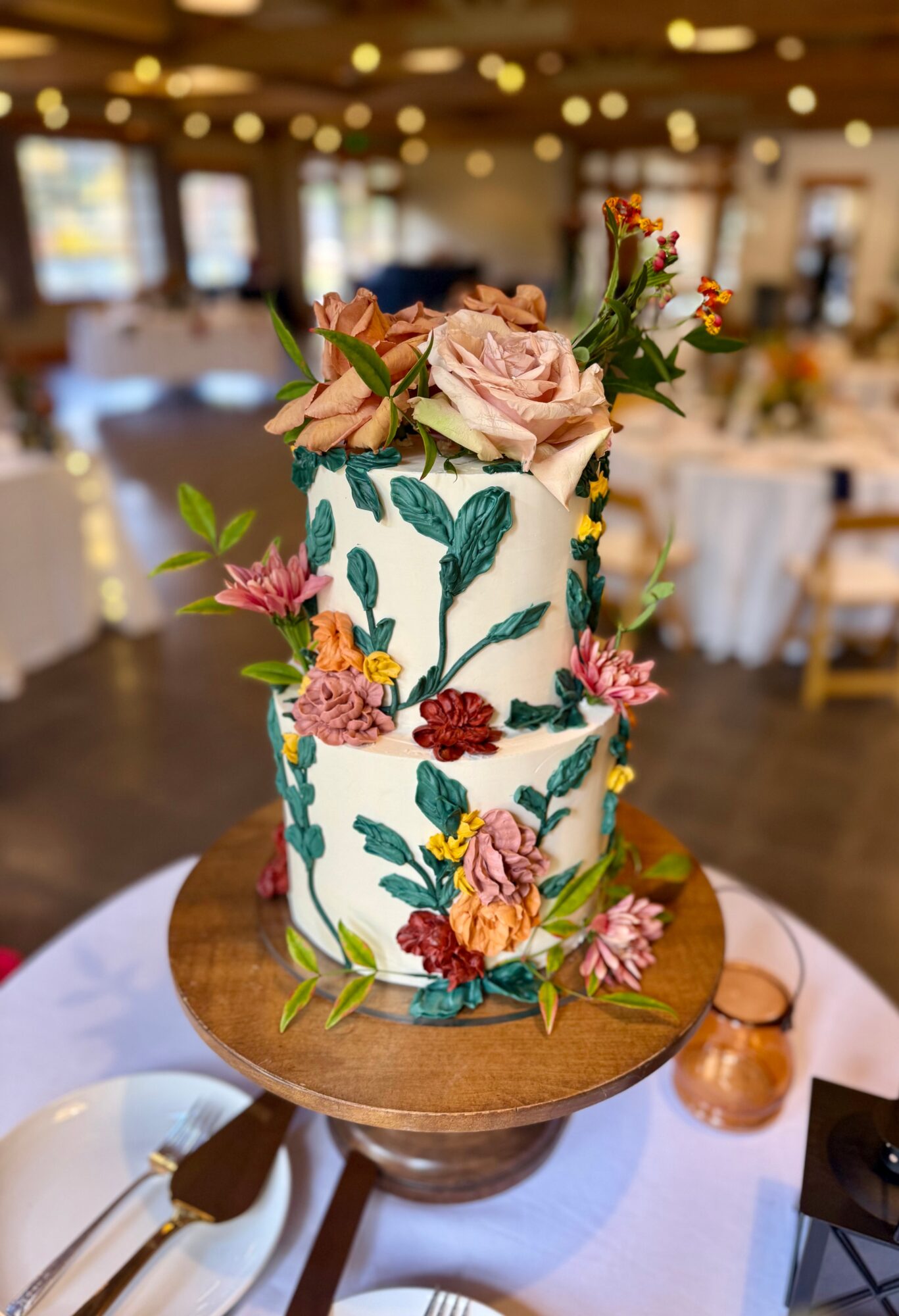 Two-tiered wedding cake decorated with pink, red, and yellow flowers and green leaves, on a wooden stand in a decorated venue.