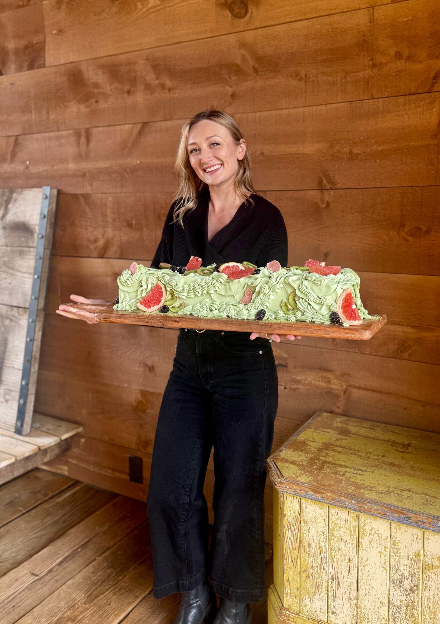 Woman holding a large decorated cake, smiling, standing in front of wooden wall, with a yellow table nearby.