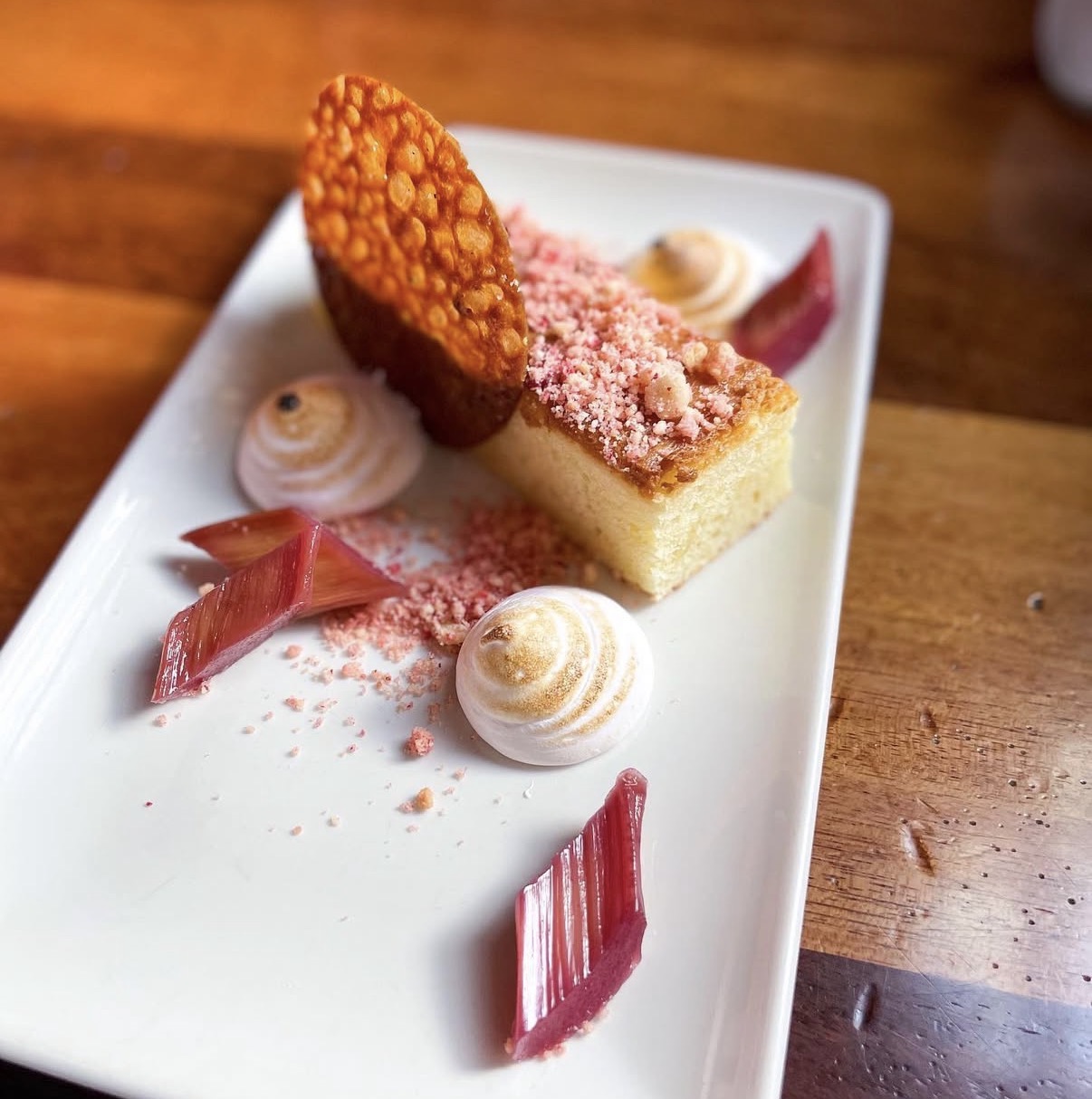 Dessert with sponge cake, pink powder, meringue, and pink decorative pieces on a white rectangular plate, on a wooden table.