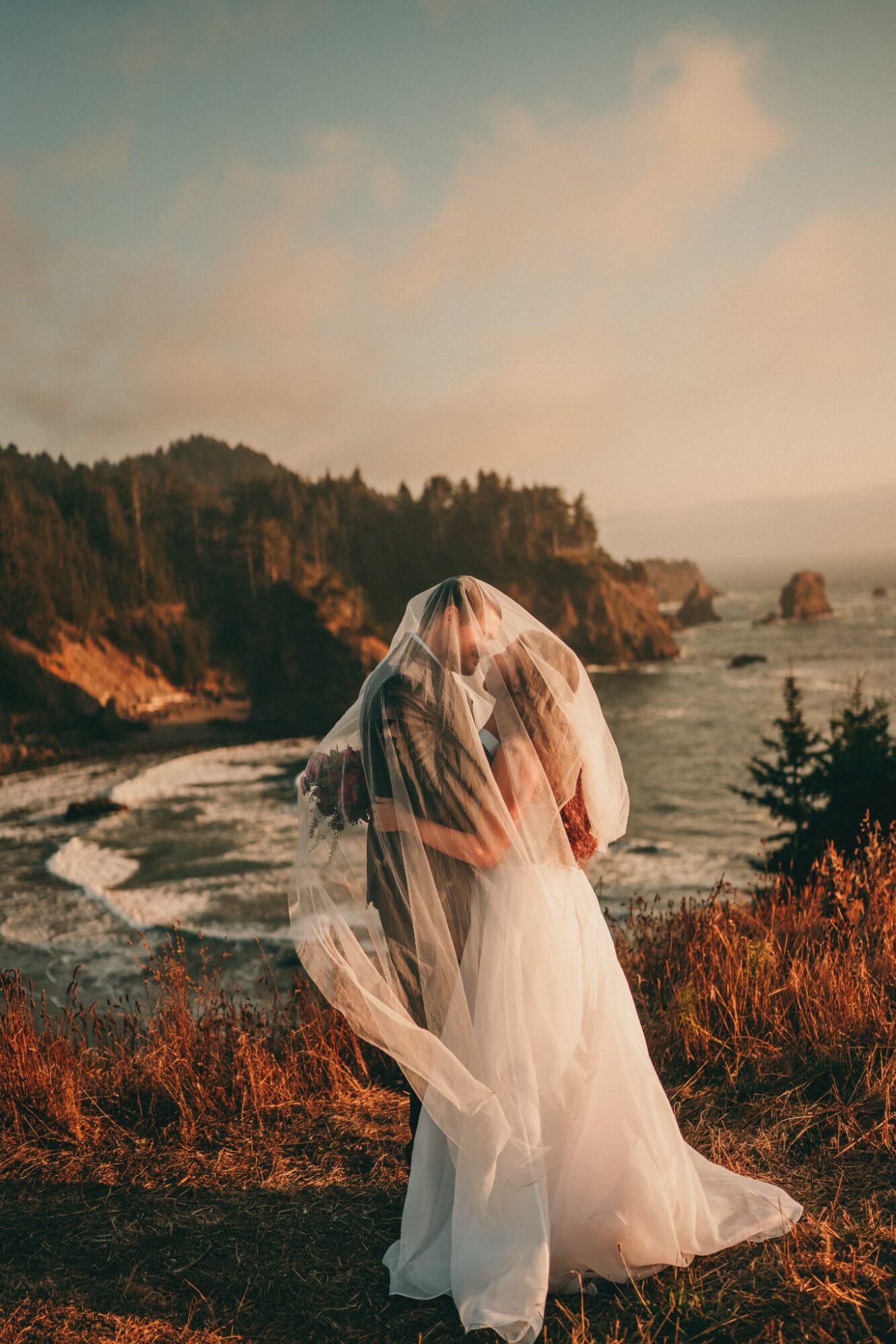 Person in wedding dress and veil standing outdoors near coastline with trees and ocean in background.