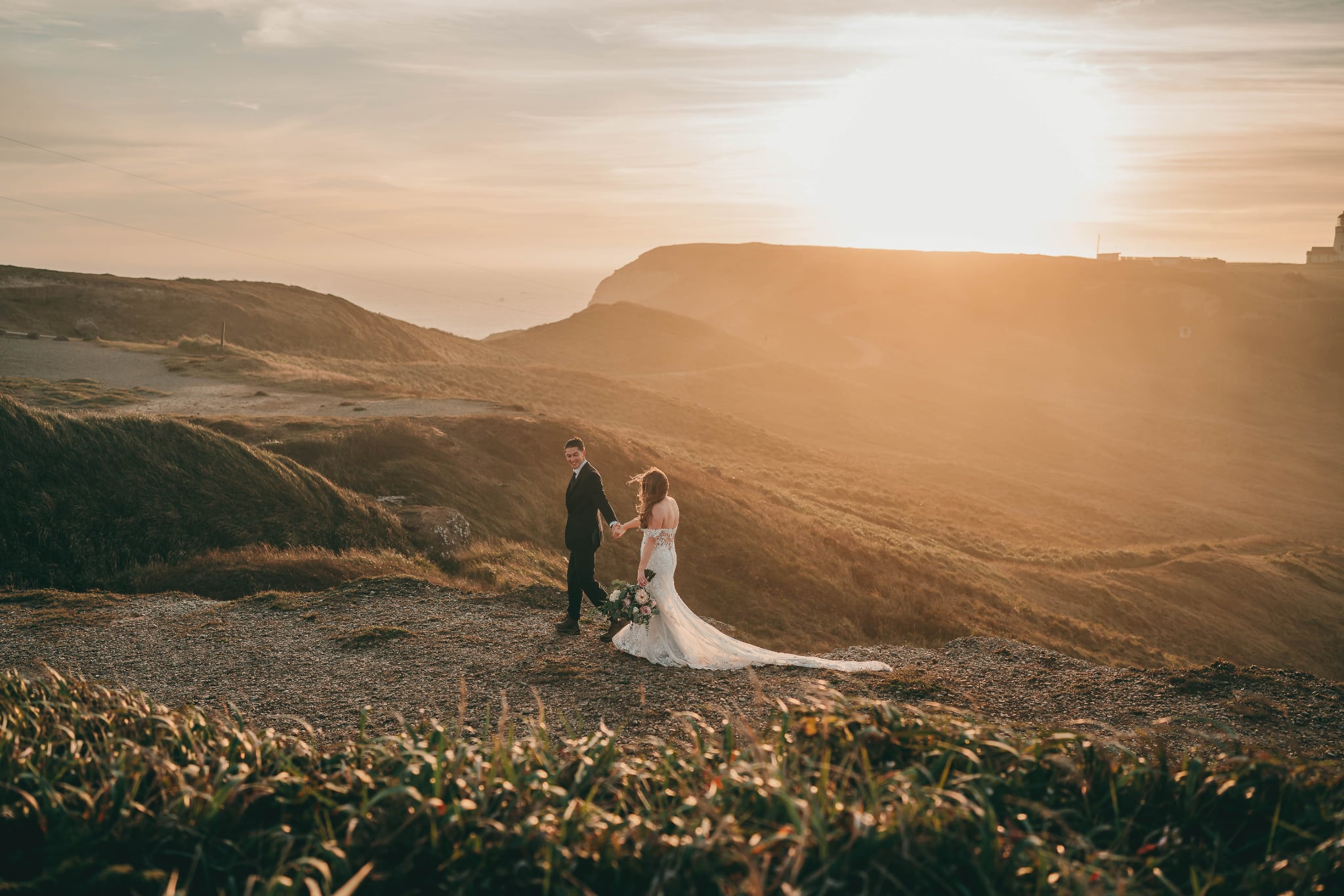 A couple stands on a hill at sunset, with a vast landscape and sky in the background.