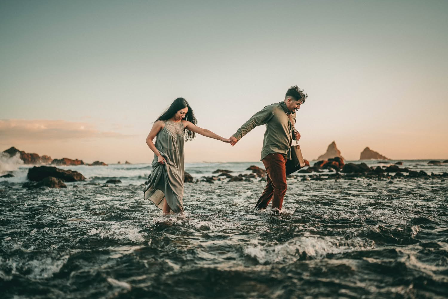 Two people holding hands walking through shallow ocean water at sunset, rocks in the background, sky clear.
