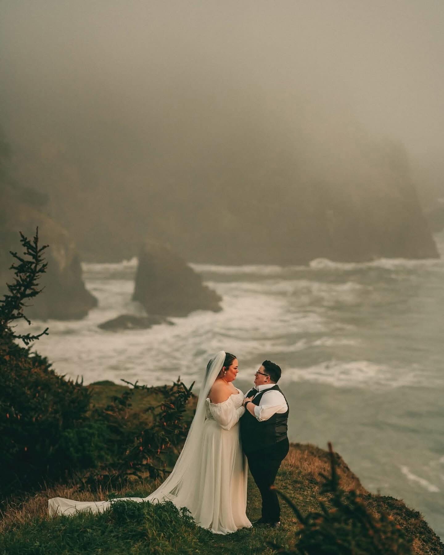 A bride and groom stand on a rocky coastline with ocean waves and cliffs in the background.