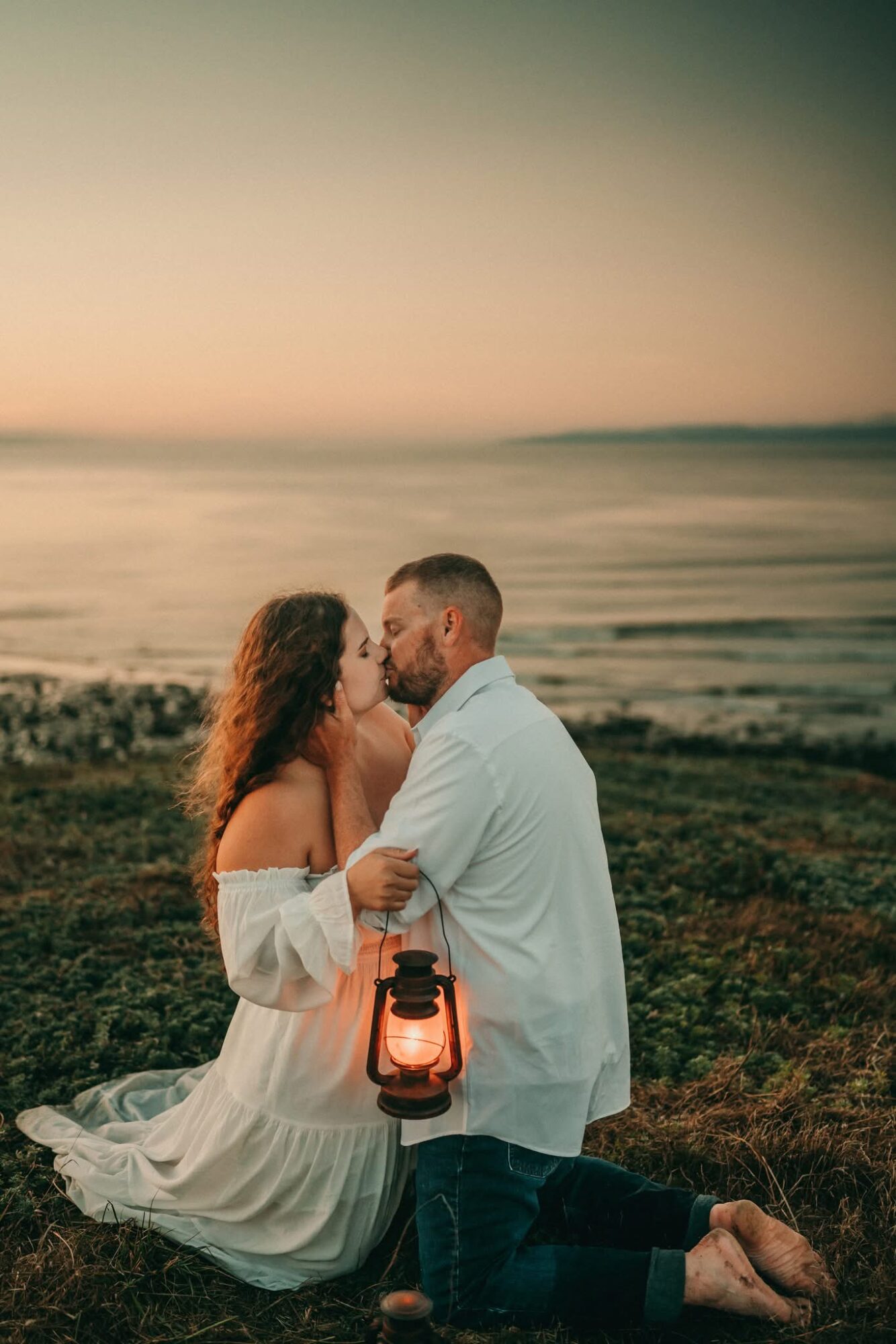Couple sitting on grass near the ocean at sunset, embracing and about to kiss, with a lantern beside them.
