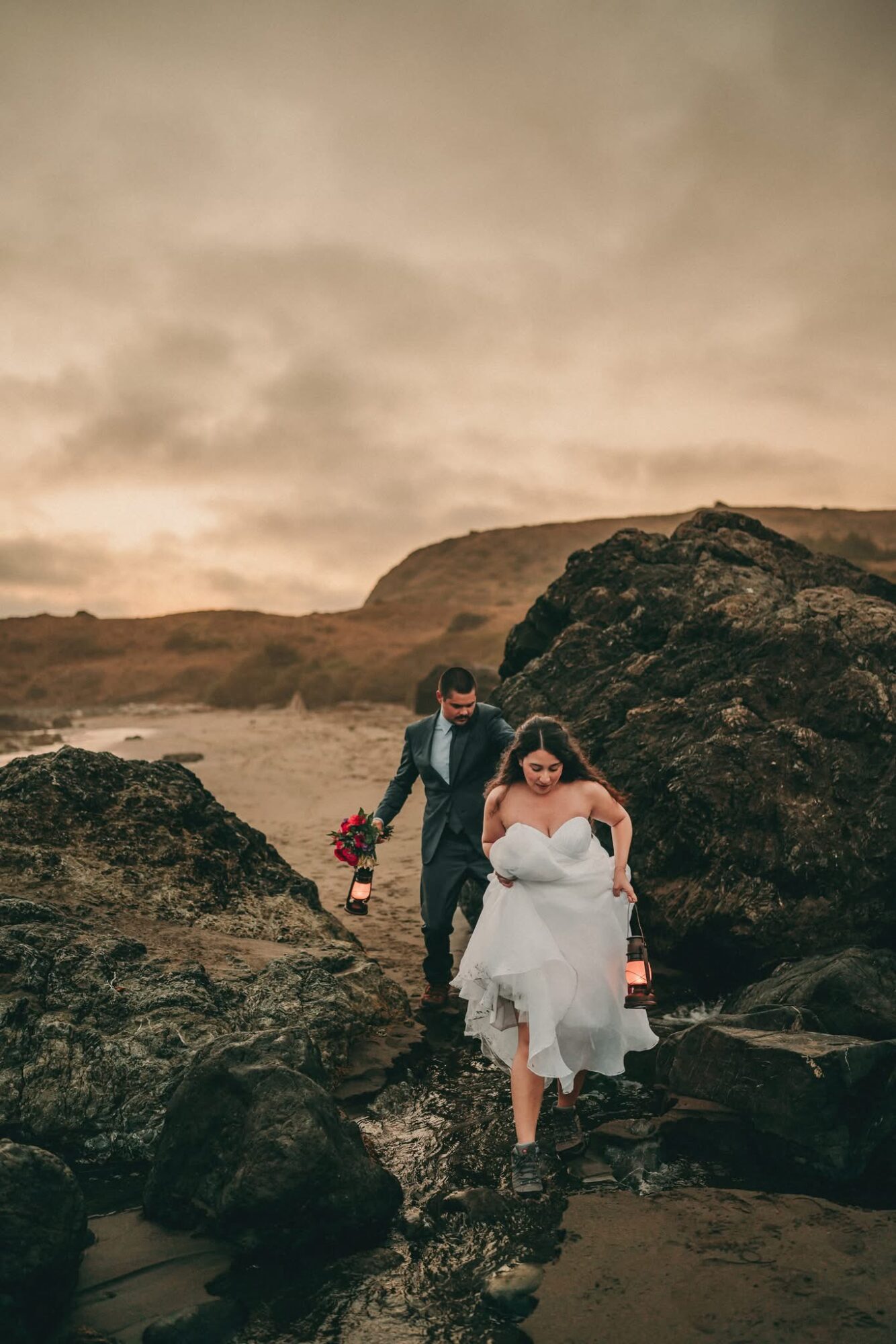A couple walking on rocks at the beach, with a cloudy sky and cliffs in the background, the woman in a white dress.