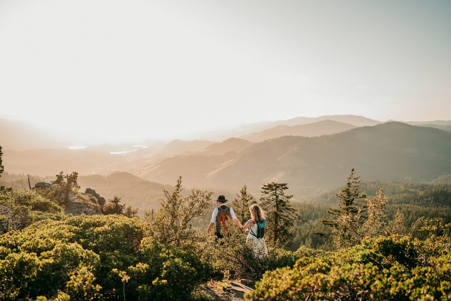Two people walk on a trail through lush green trees with mountains in the background, under a bright sky.