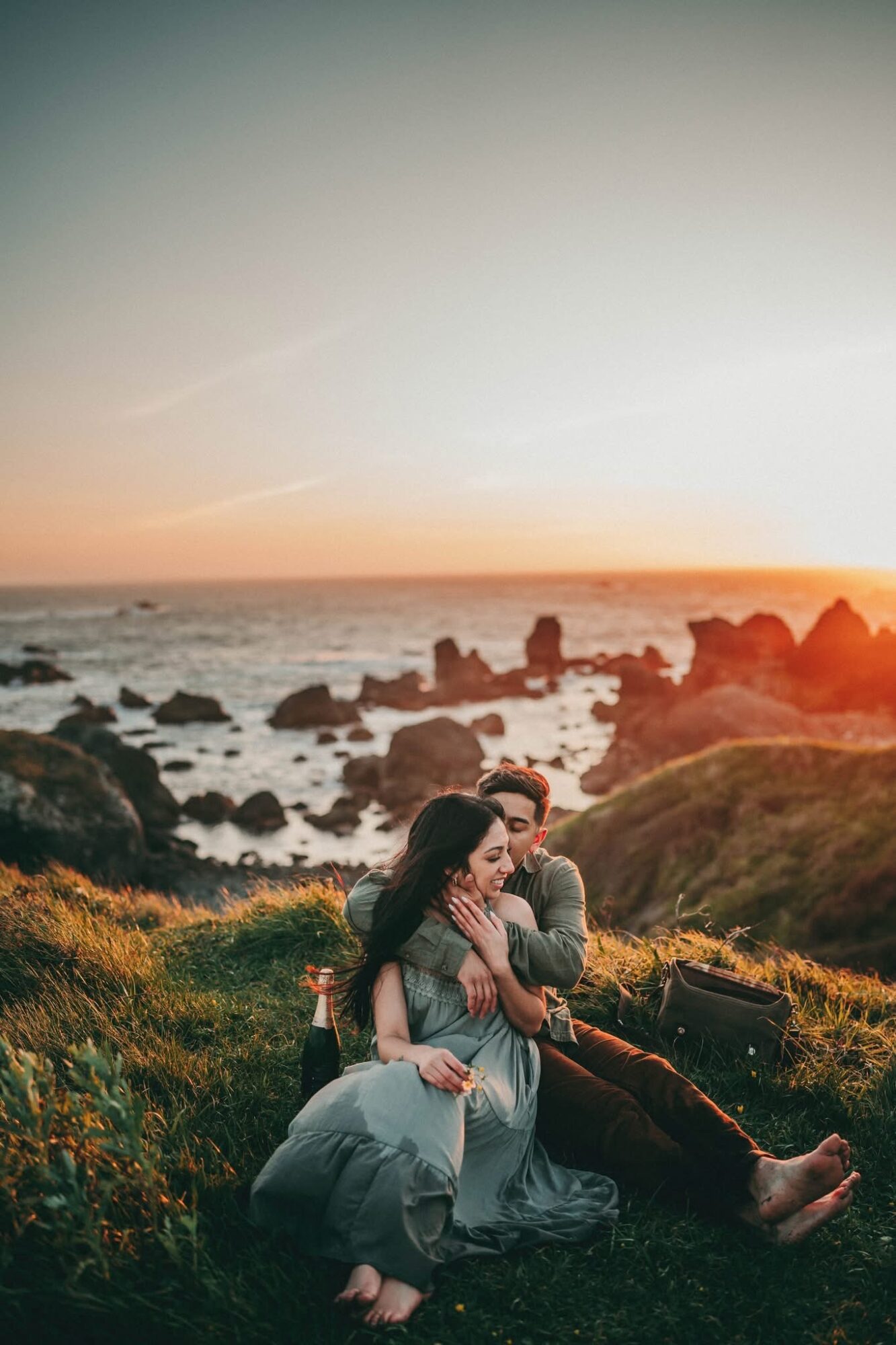 Two people sit on rocks near the ocean at sunset, embracing with a rocky shoreline and sky in the background.