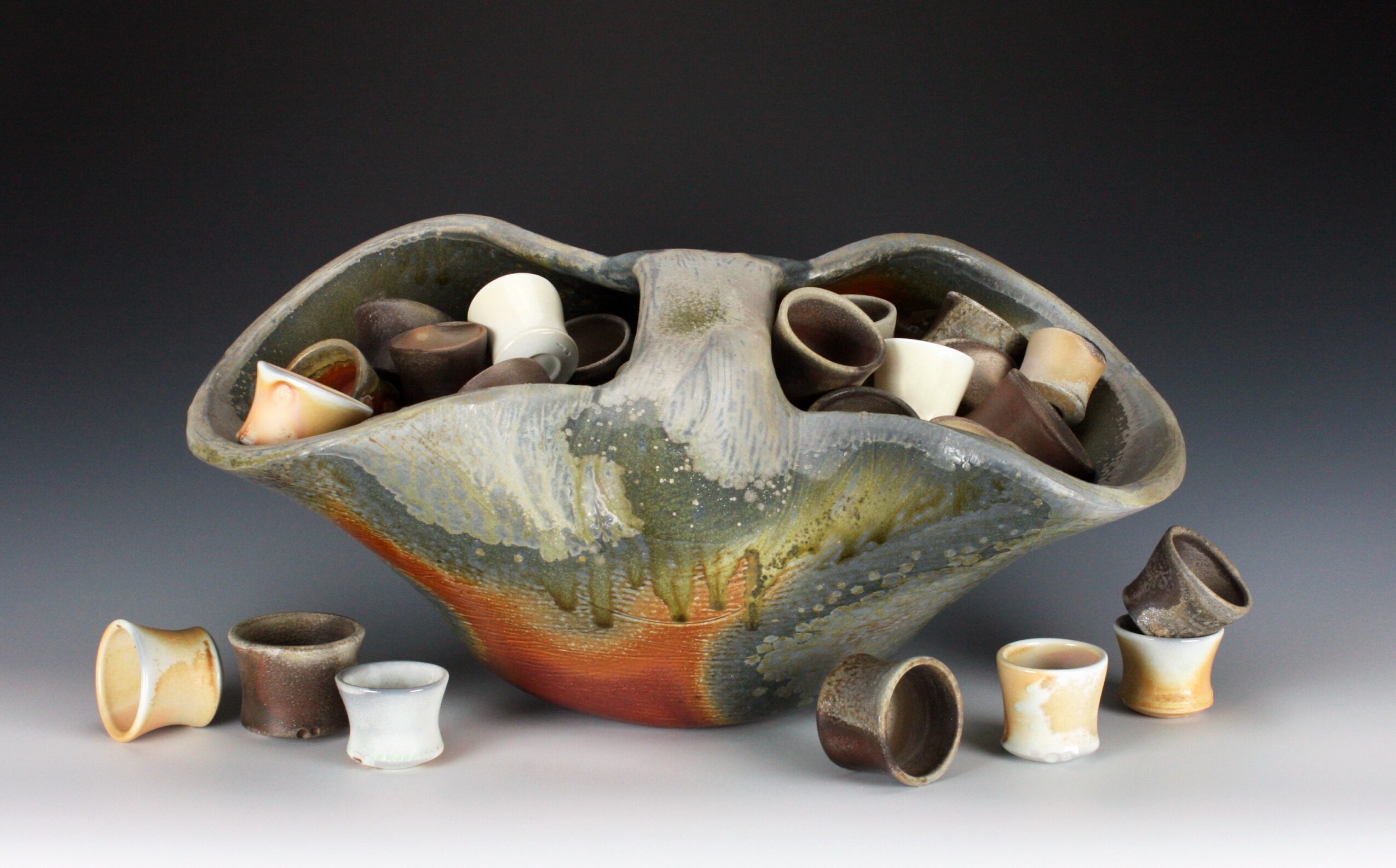 Decorative ceramic bowl filled with small, colorful cups, some outside the bowl, on a dark background.