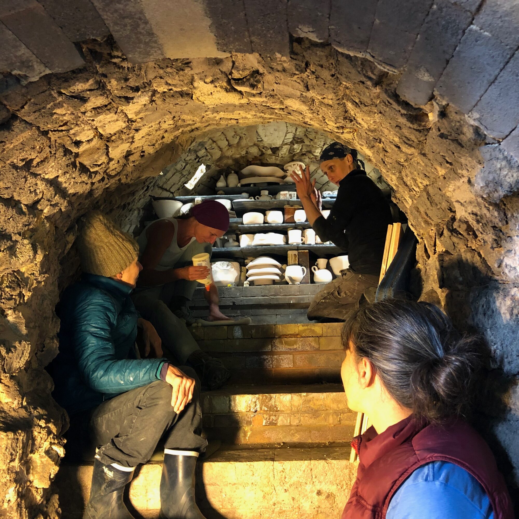 People sitting and looking at shelves with dishes inside a stone archway. One person is taking a photo. Bright lighting.