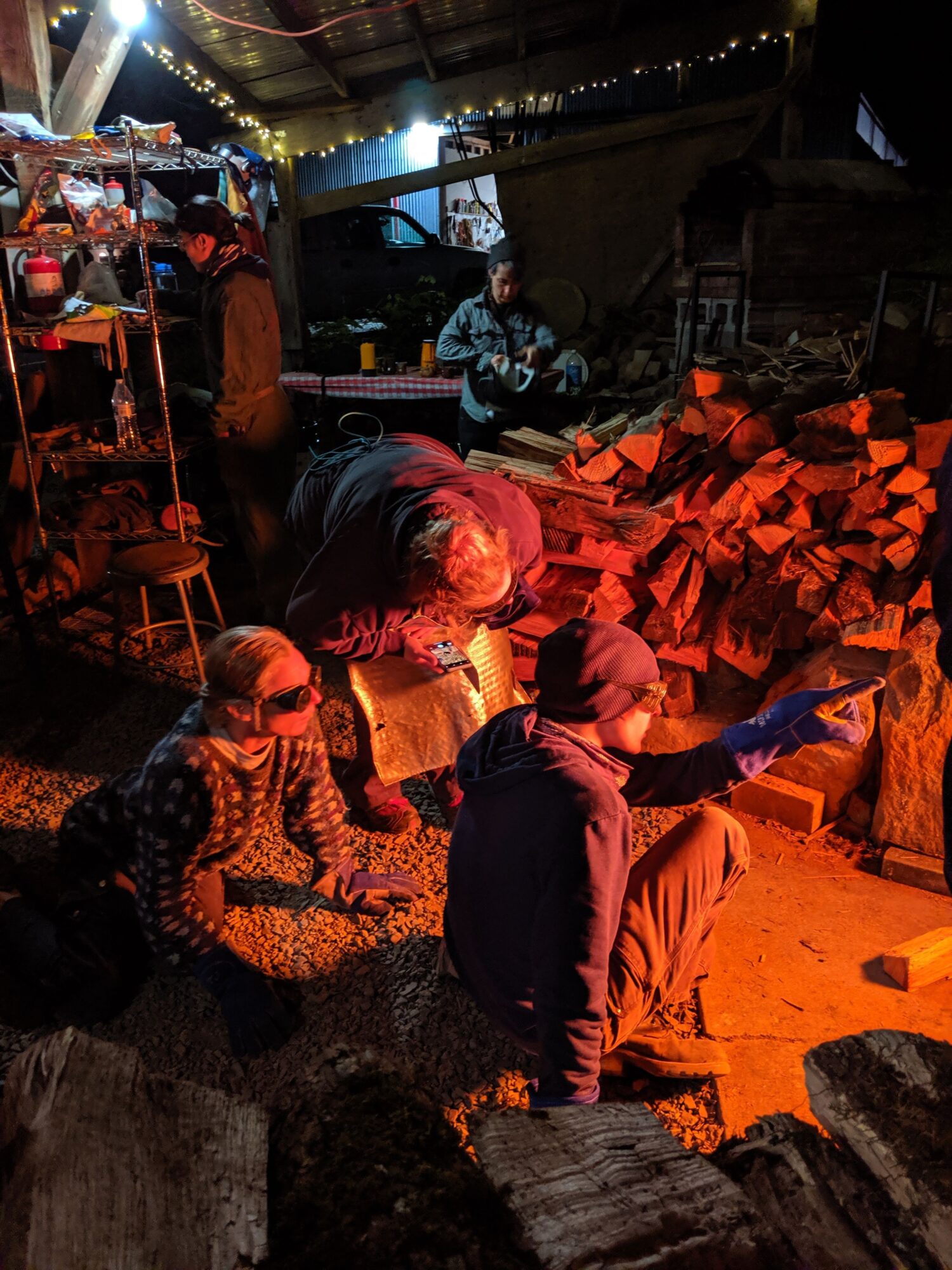 People sitting and talking around a fire pit outdoors at night, with stacked firewood in the background.