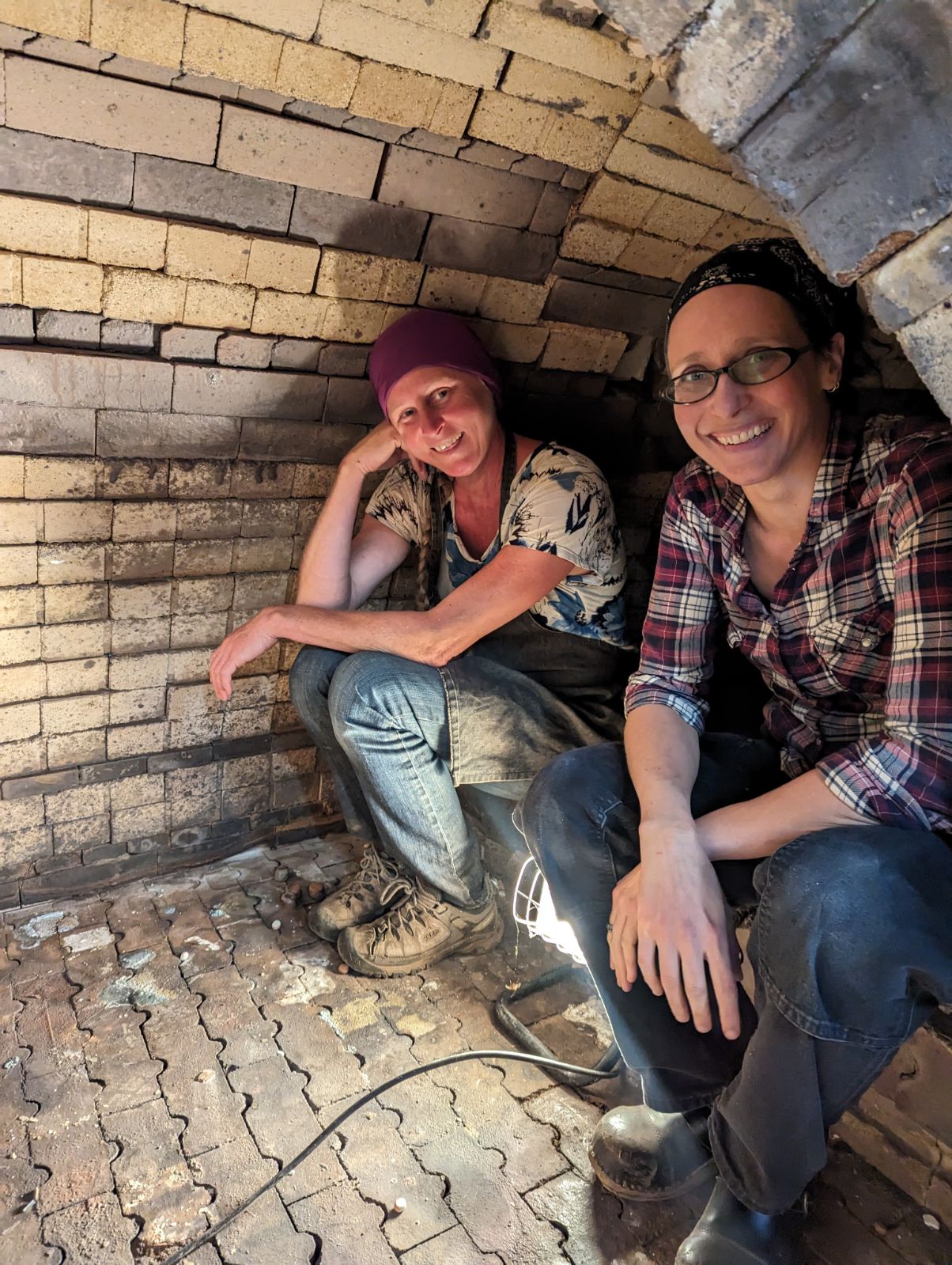 Two people sitting inside a brick archway, smiling, one wearing glasses and a plaid shirt, the other with a beanie and casual clothing.