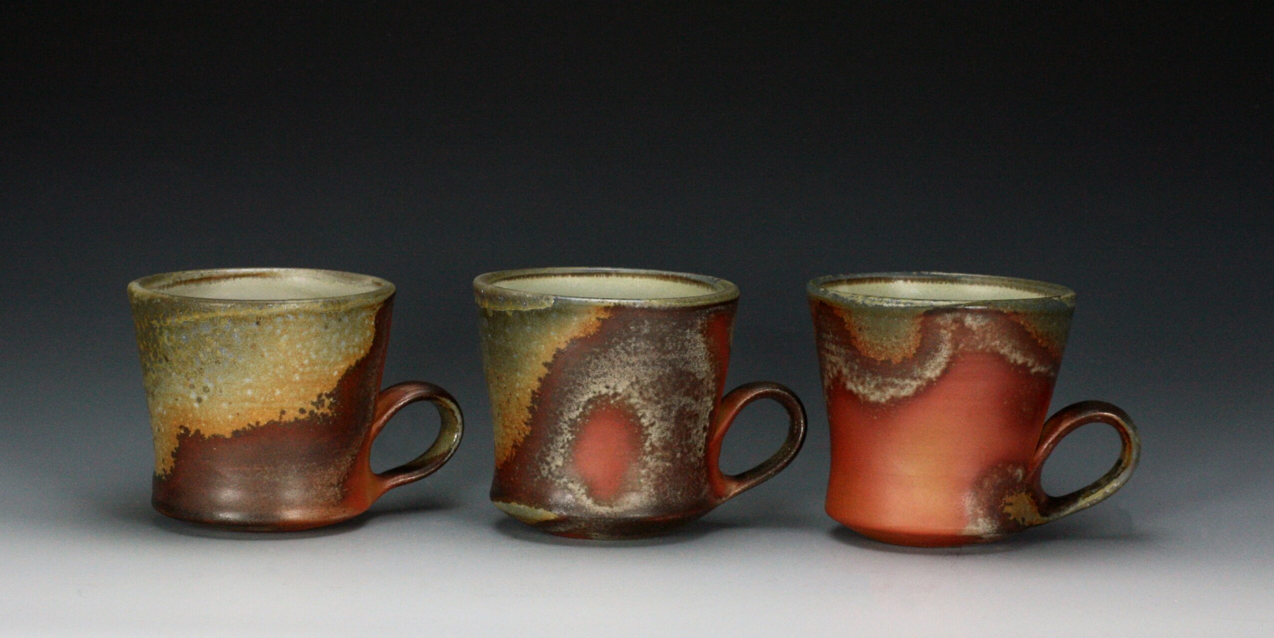 Three ceramic cups with abstract glaze patterns, arranged in a row against a dark background.