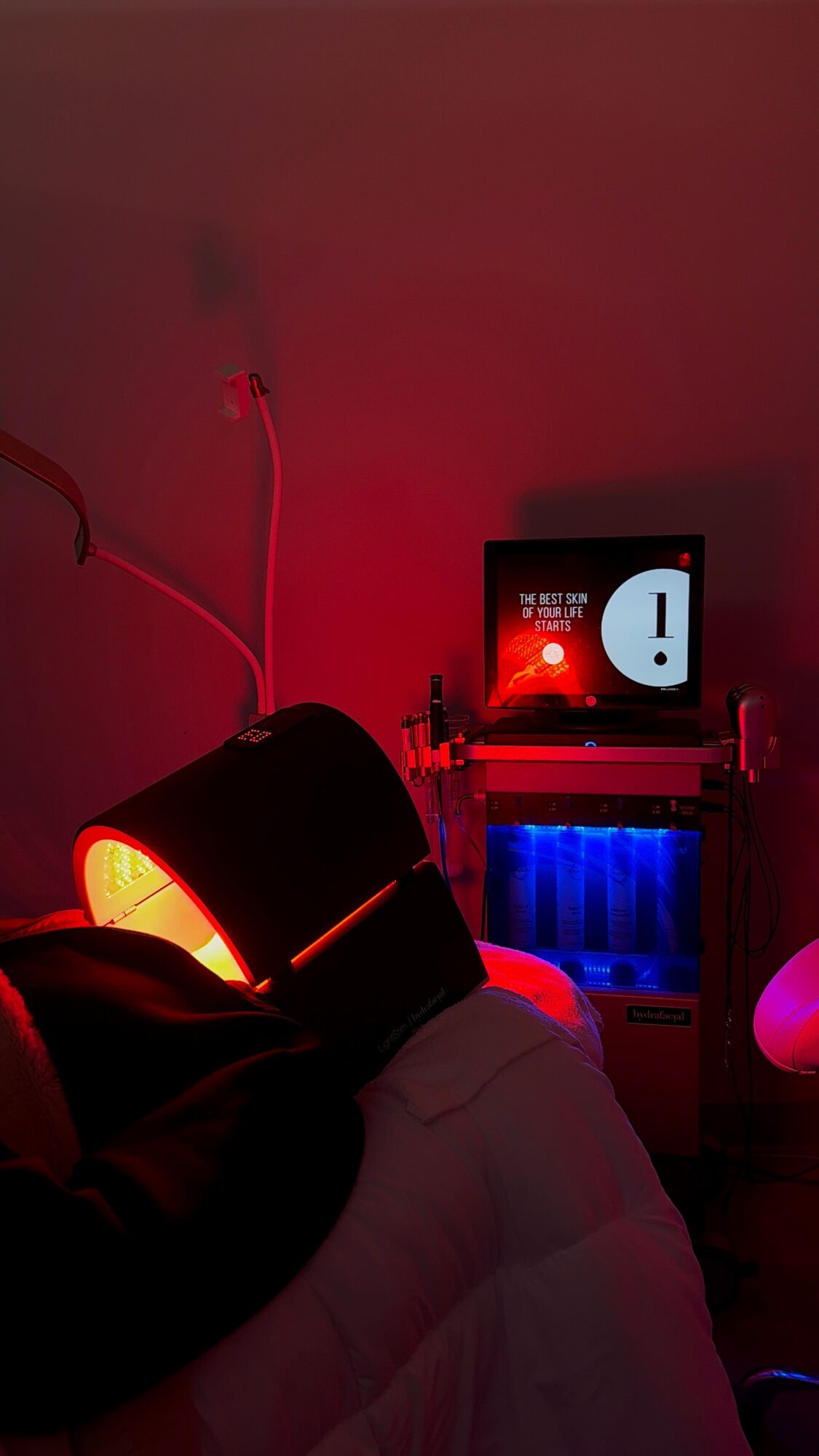 A dimly lit room with a person lying on a bed, a small monitor displaying an exclamation mark, and colorful lighting.
