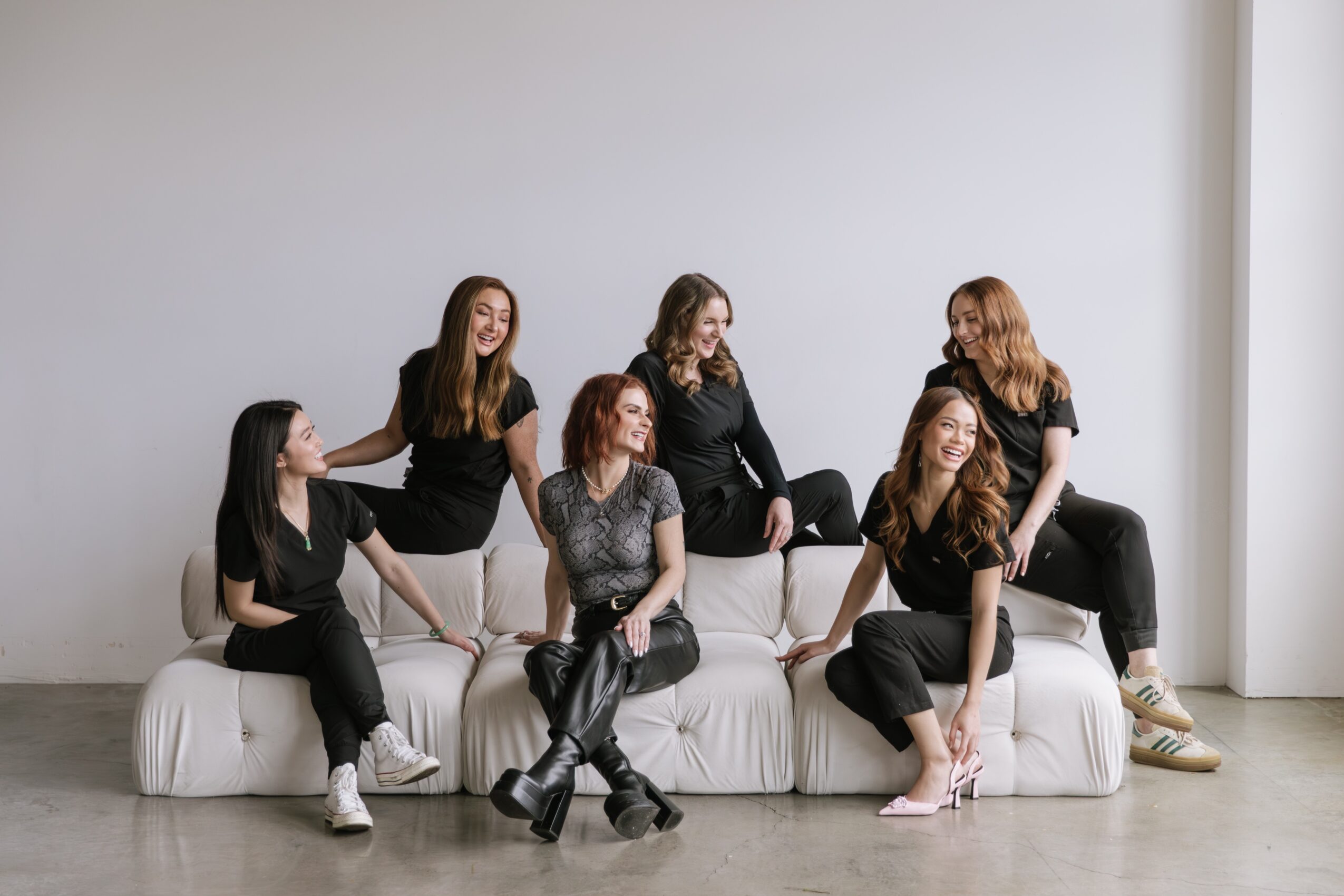 Six women sitting and standing on a white sofa in a minimal room, smiling and interacting with each other.
