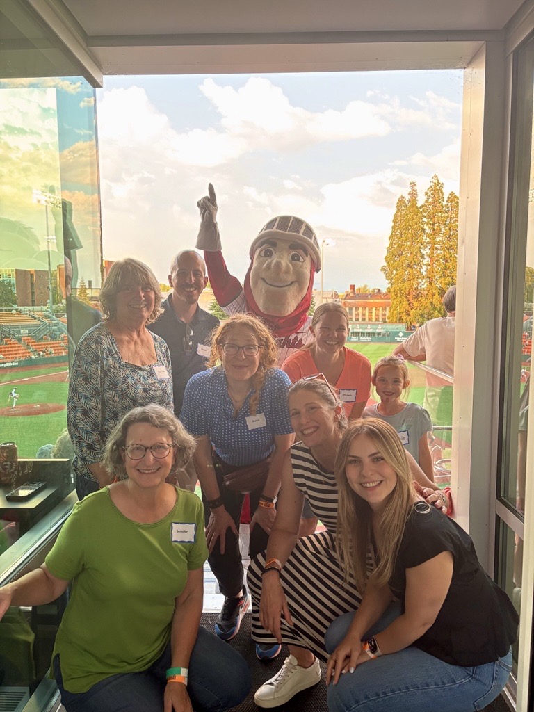 Group of people, including a person in a mascot costume, standing inside near a window with outdoor view, smiling.