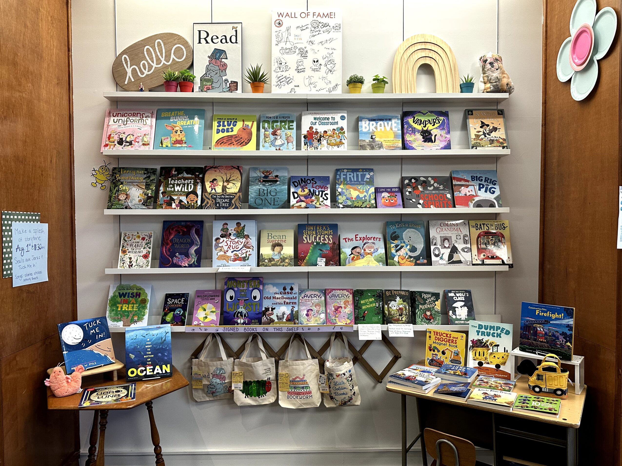 Bookshelves with children's books, decorations, and signs in a cozy reading area, with a small table displaying toys and books.