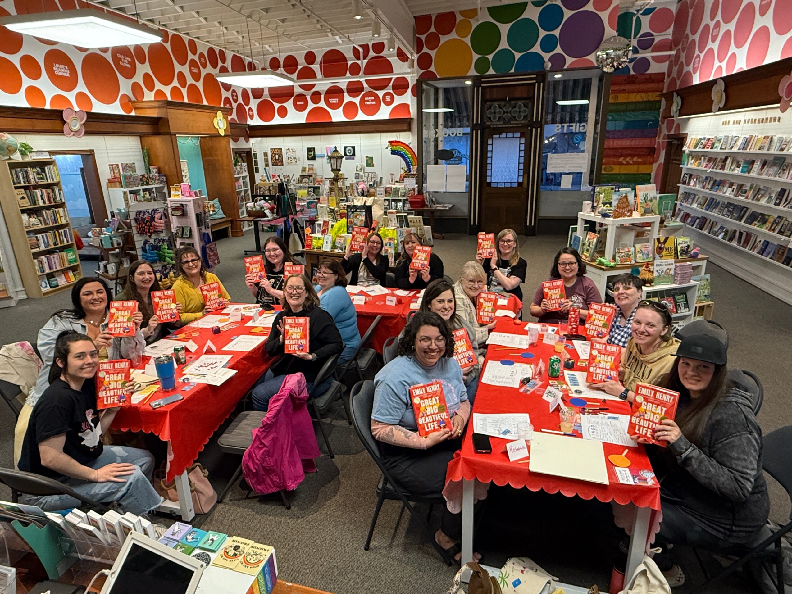 Group of people sitting at red-covered tables in a colorful bookstore, holding books and smiling.