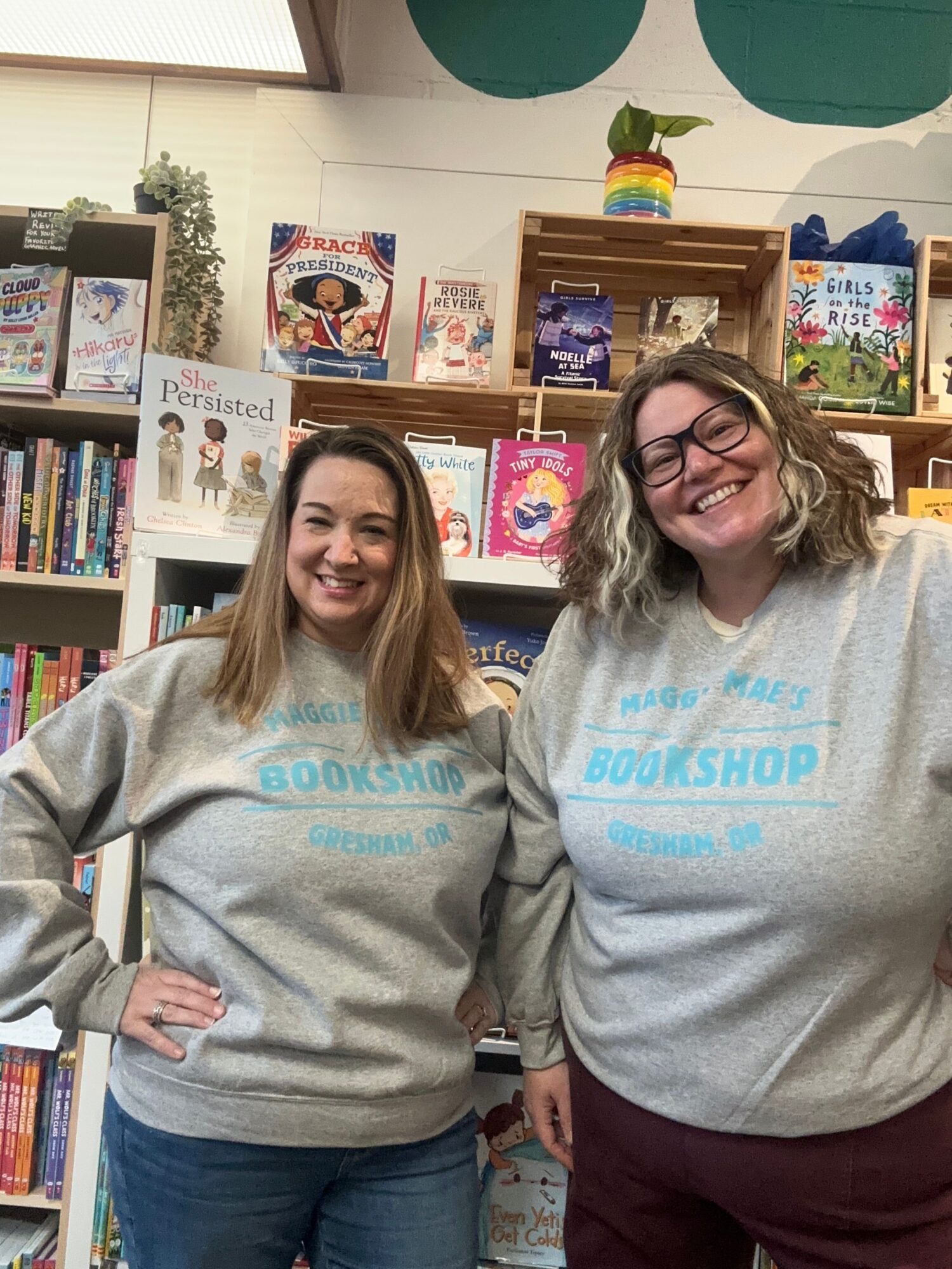 Two women standing in a bookstore, smiling, with bookshelves and colorful books in the background.