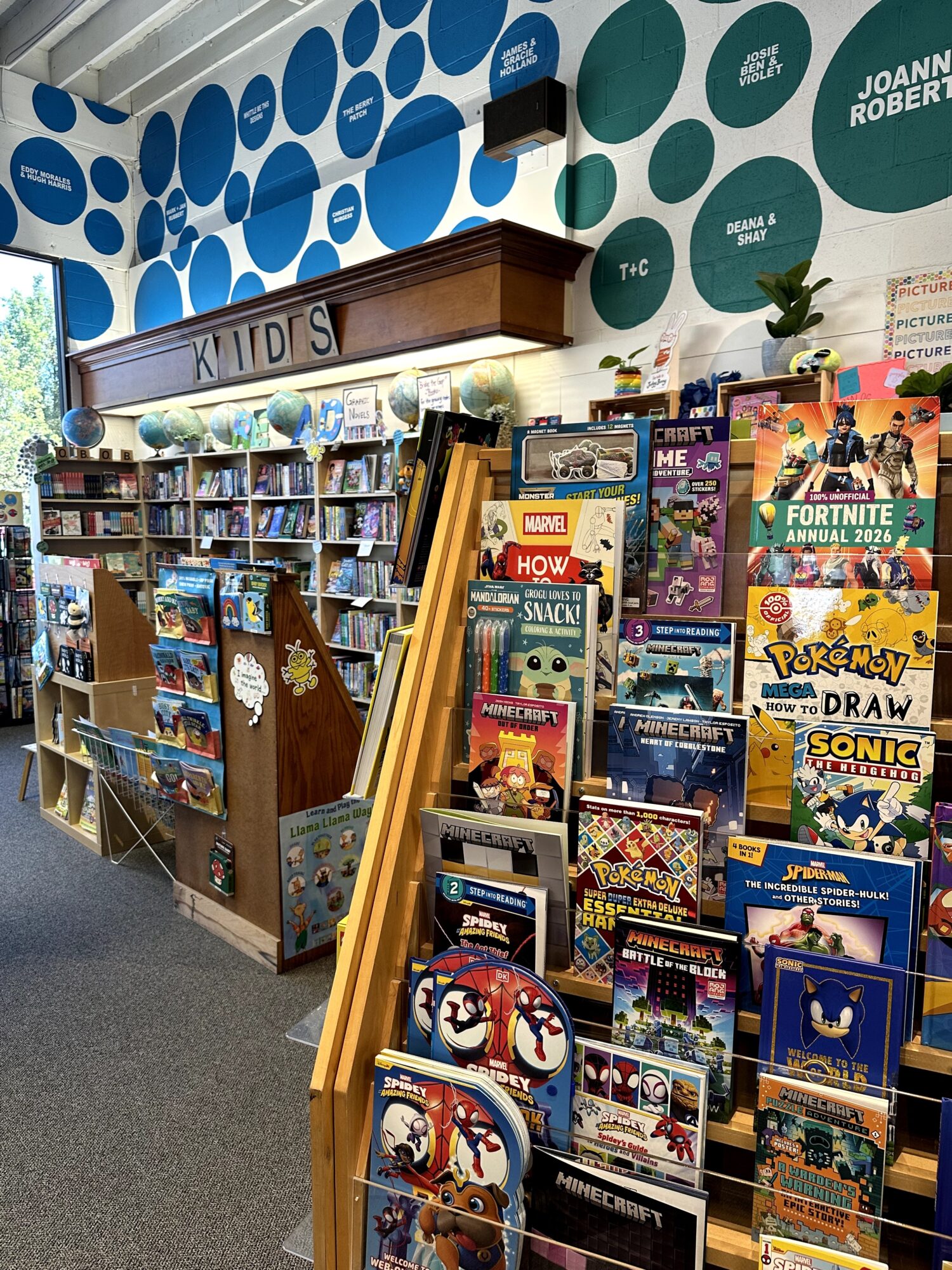 Bookshelf filled with children's books and comics, with a colorful wall of circles and a window in the background.