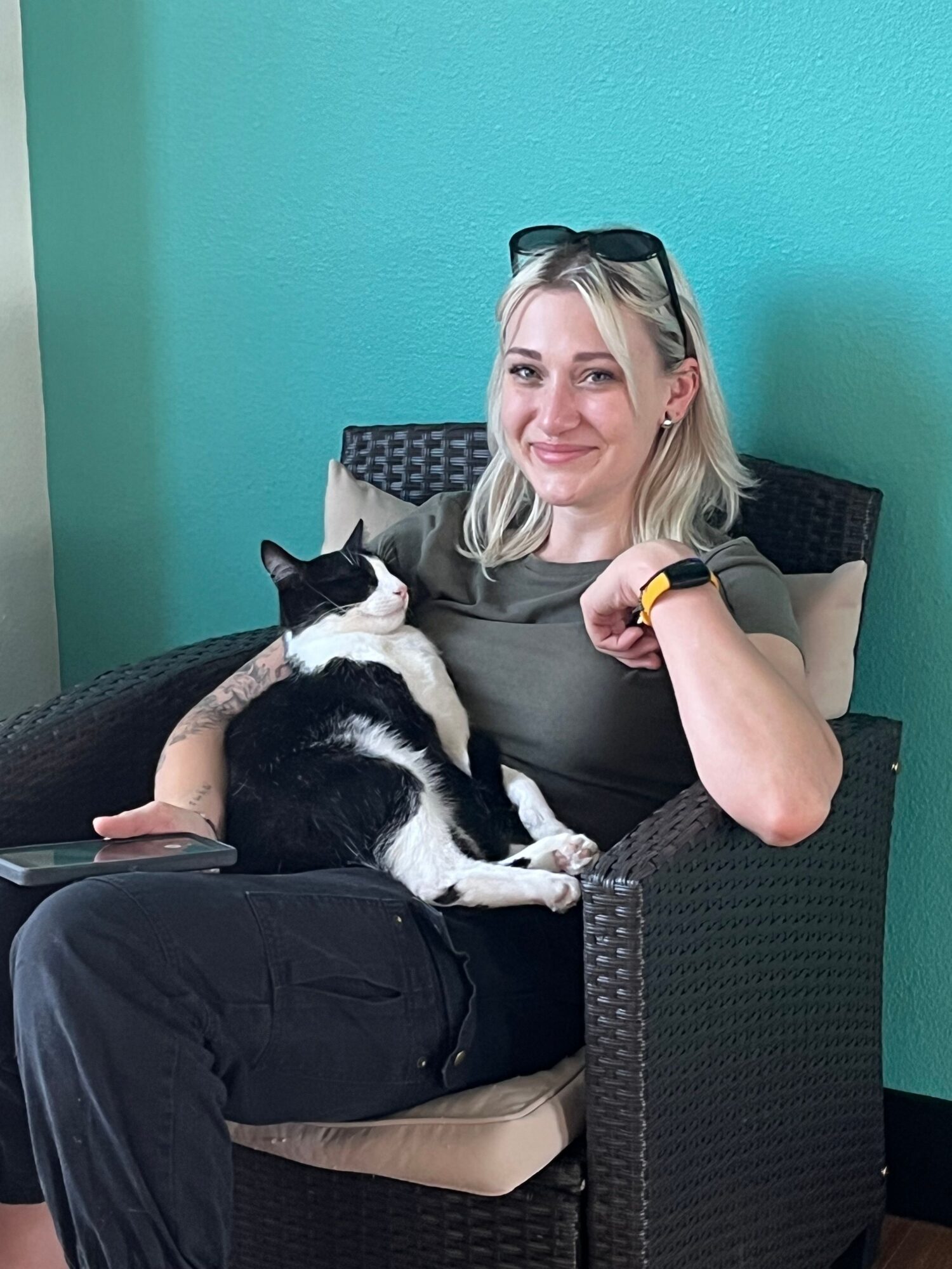 Woman sitting on a black outdoor chair with a black and white cat on her lap, against a teal wall.