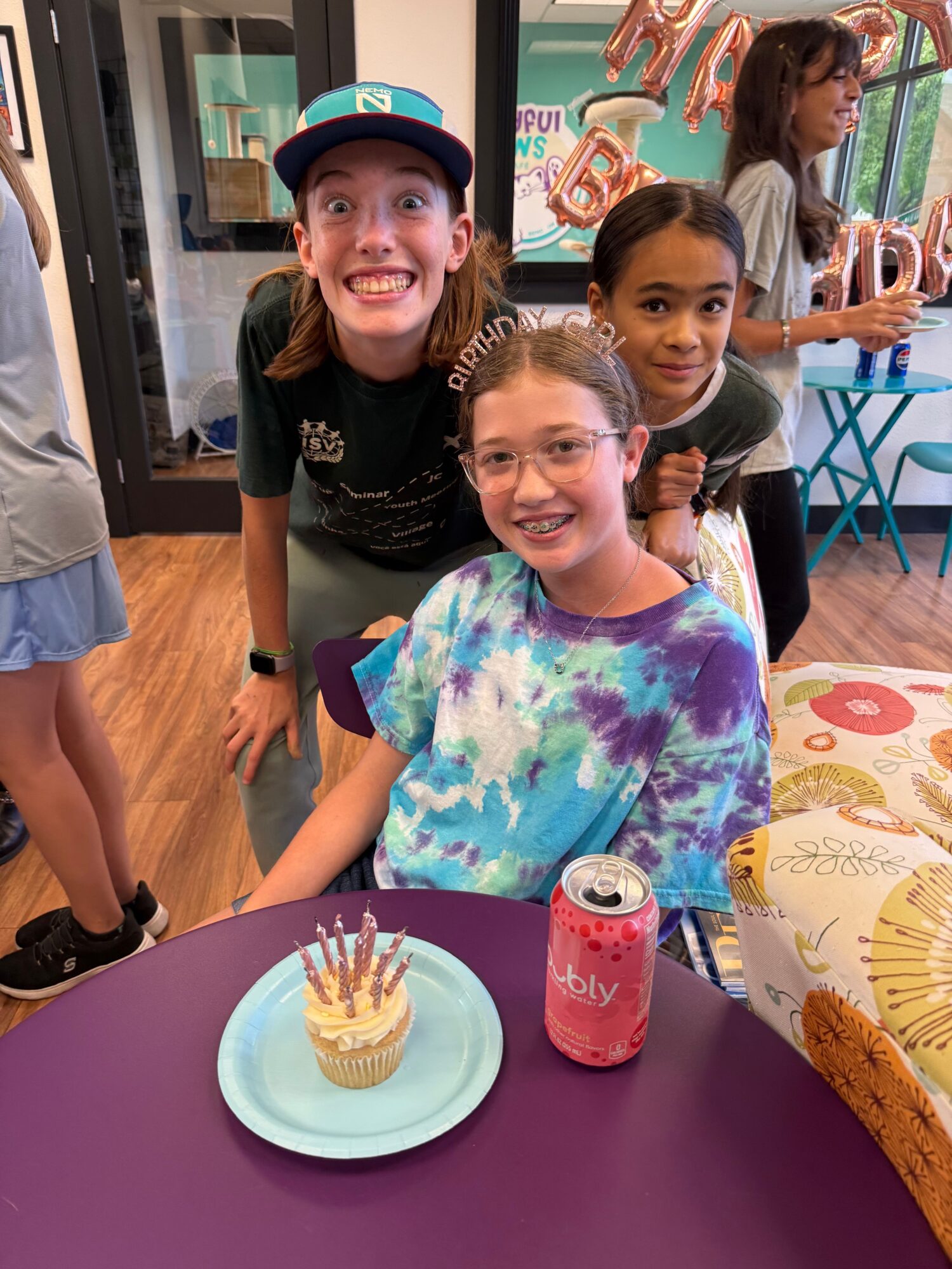 Three women smiling at a birthday celebration, with a cake and drinks on a purple table, in a decorated room.