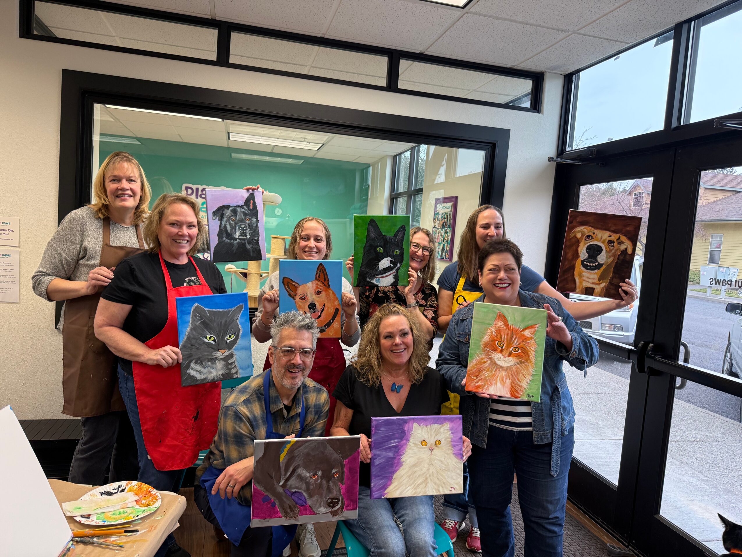 Group of nine people holding colorful animal paintings inside a room with large windows and a glass door.