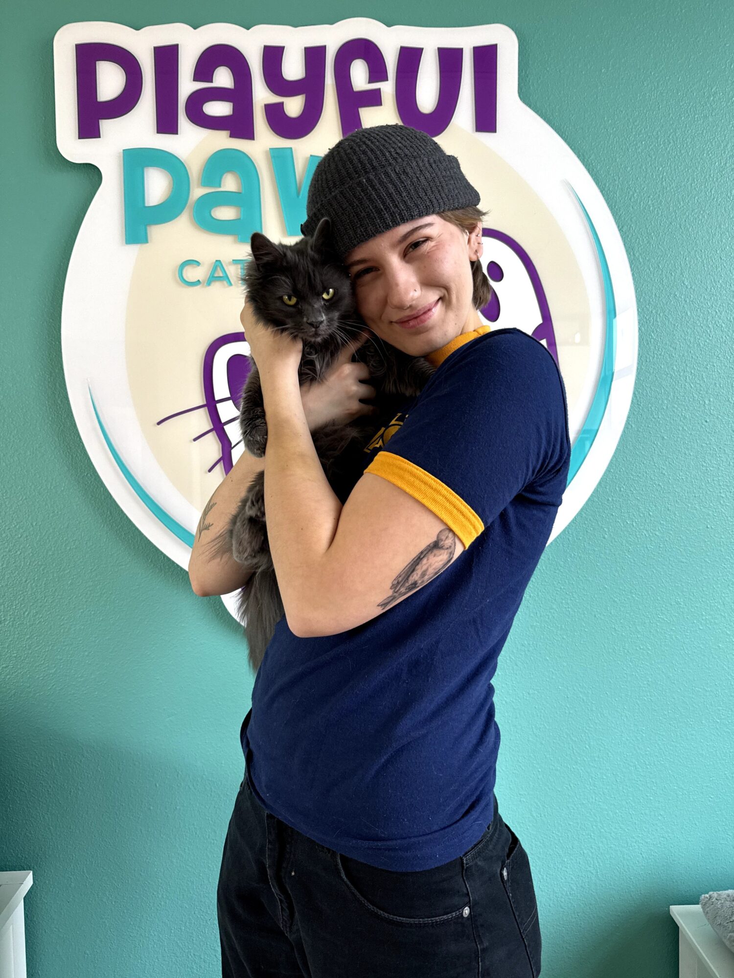 Person holding a black kitten, smiling, wearing a black beanie and blue shirt, standing in front of a colorful sign.