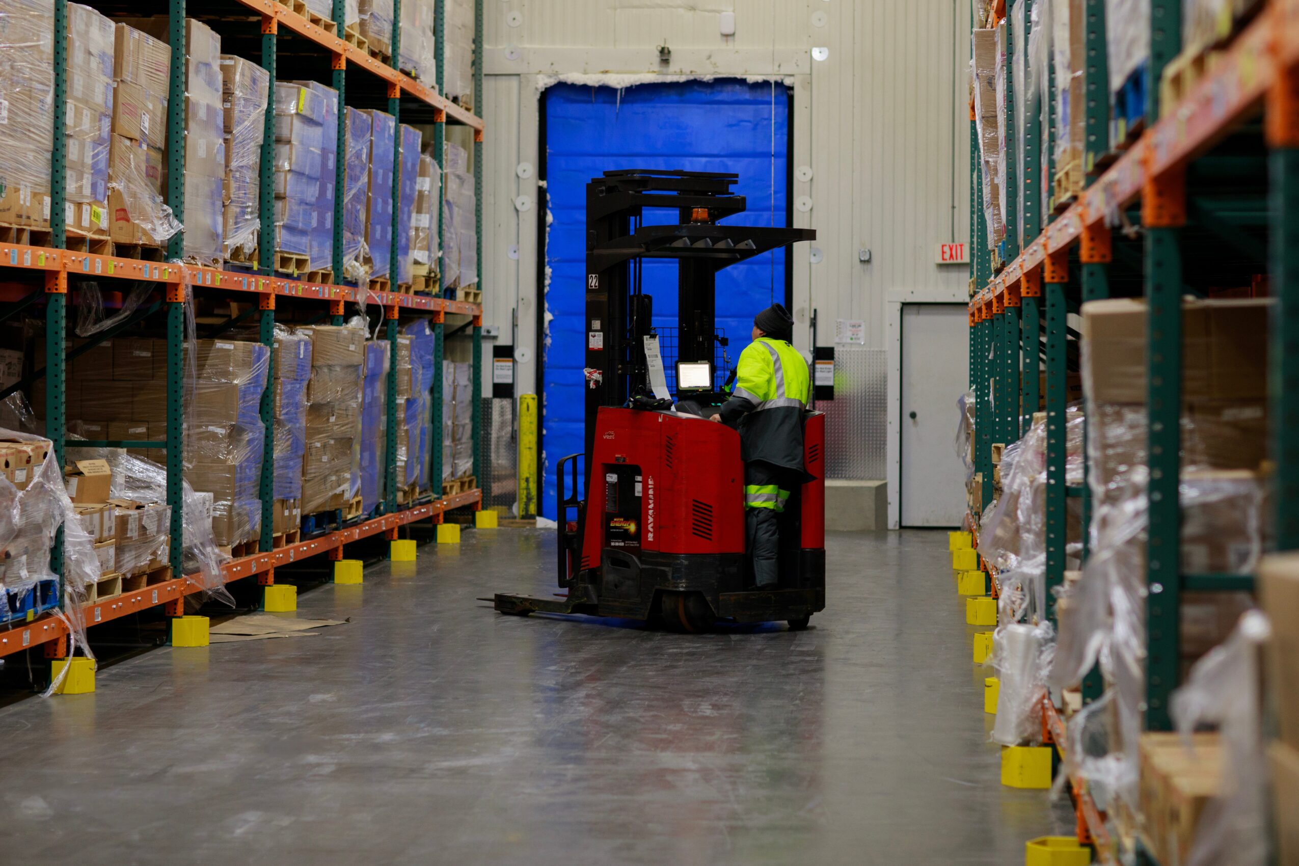 Warehouse aisle with shelves filled with boxes, a worker operating a red forklift, and a blue door in the background.