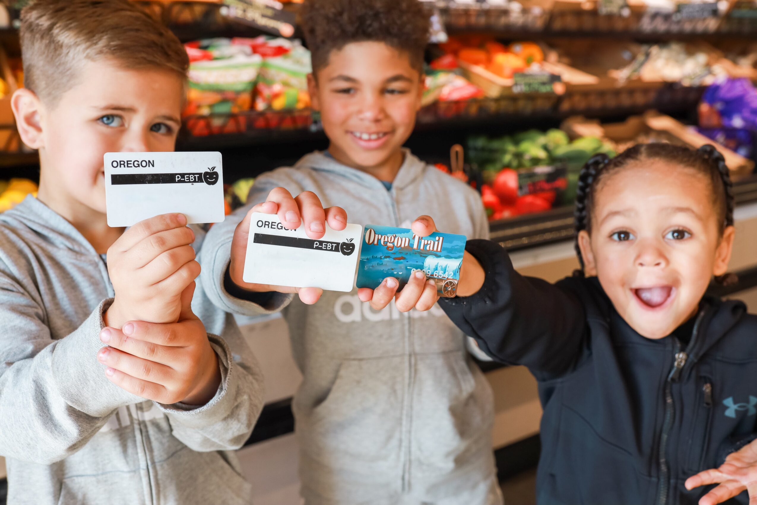 Three children smiling and holding a snack bar in a grocery store aisle with colorful produce in the background.