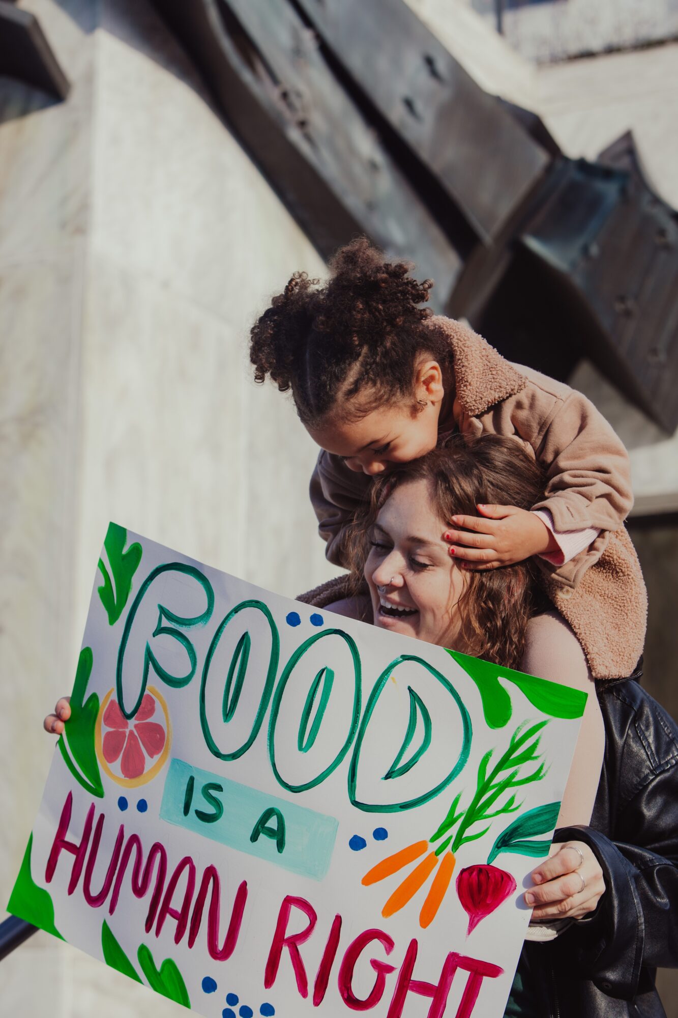 Two children, one hugging the other, holding a colorful sign that reads 'Food is a human right' with decorative leaves and hearts.